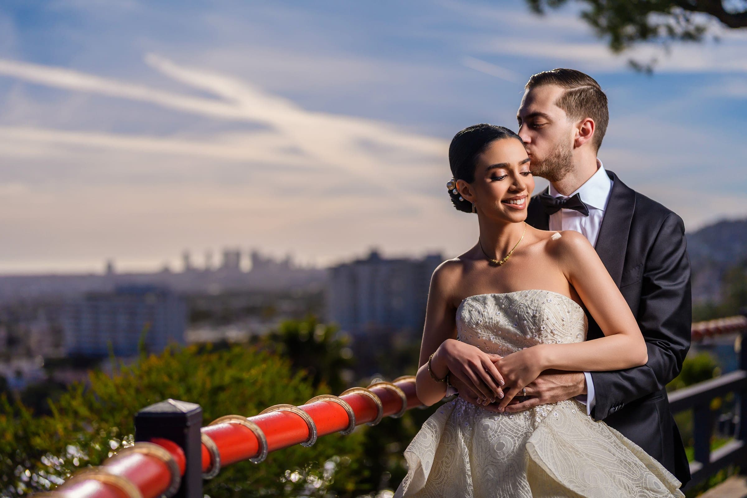 The groom kisses the bride's temple at a red railing overlooking the hazy Los Angeles skyline.