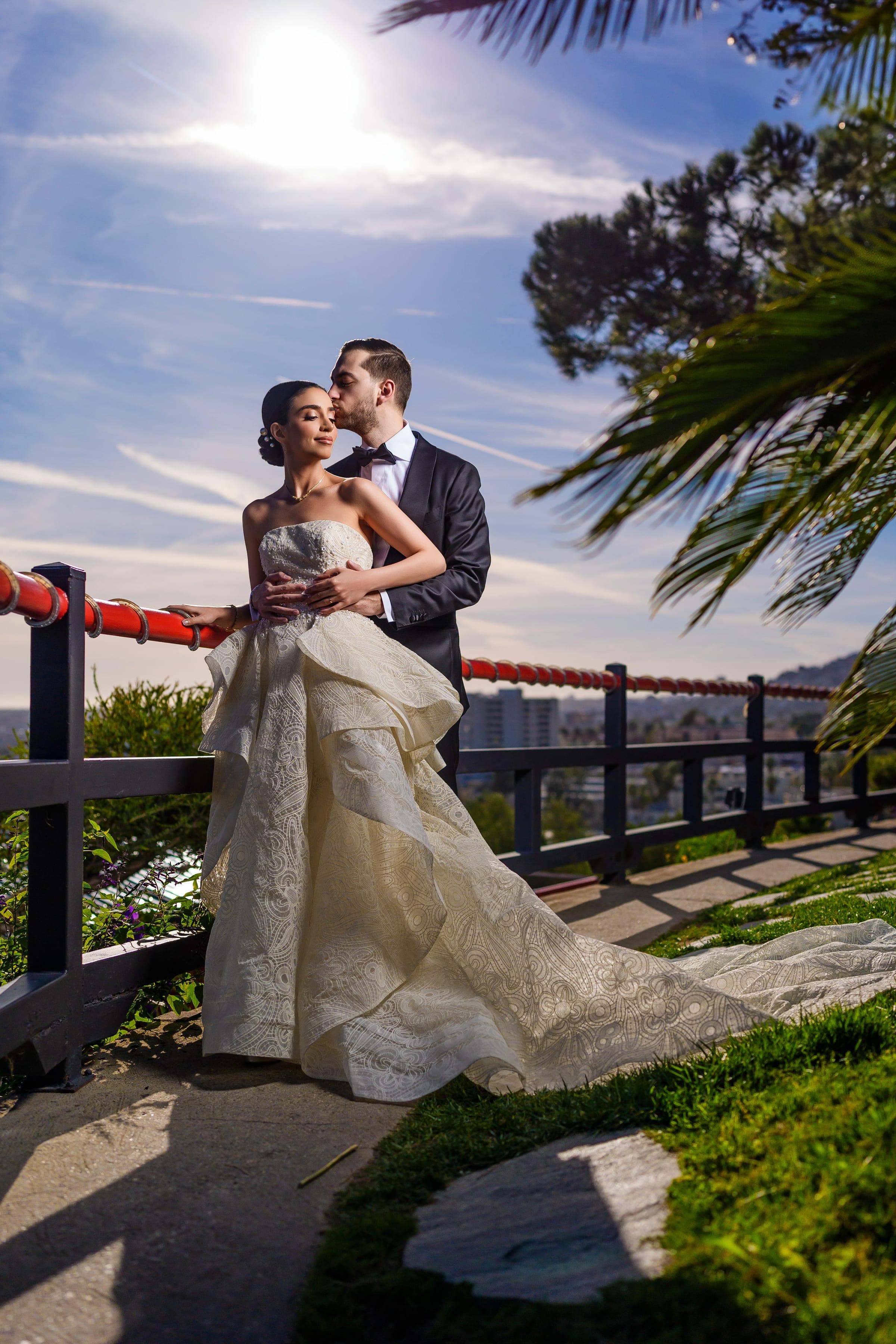 Backlit by the midday sun, the groom kisses the bride beside a red hillside railing and palm fronds.