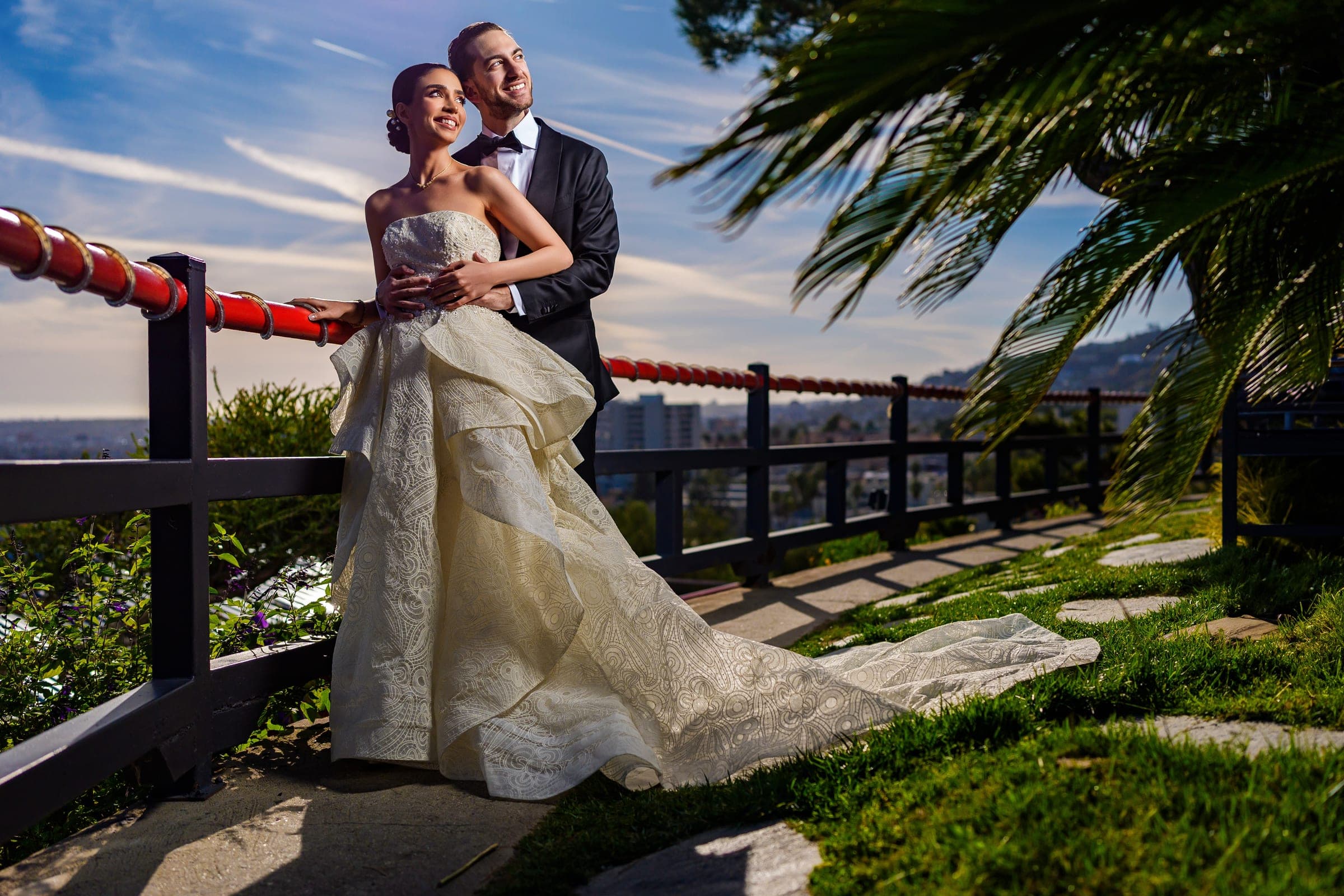 The couple smiles toward the sunlight at a red-railed overlook, her ruffled train sweeping onto the grass.