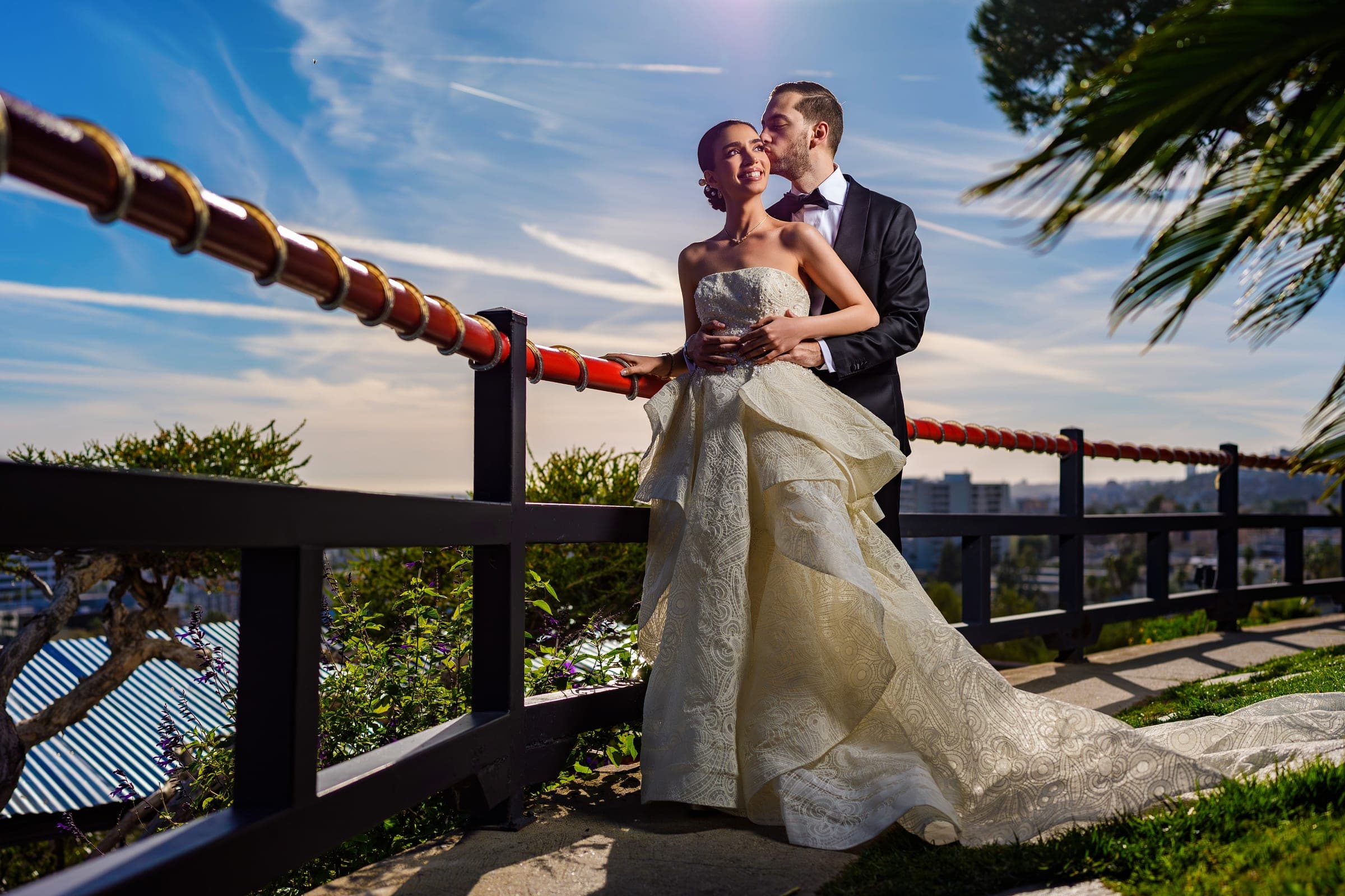The groom kisses the smiling bride against a red railing under a bright sky streaked with contrails.