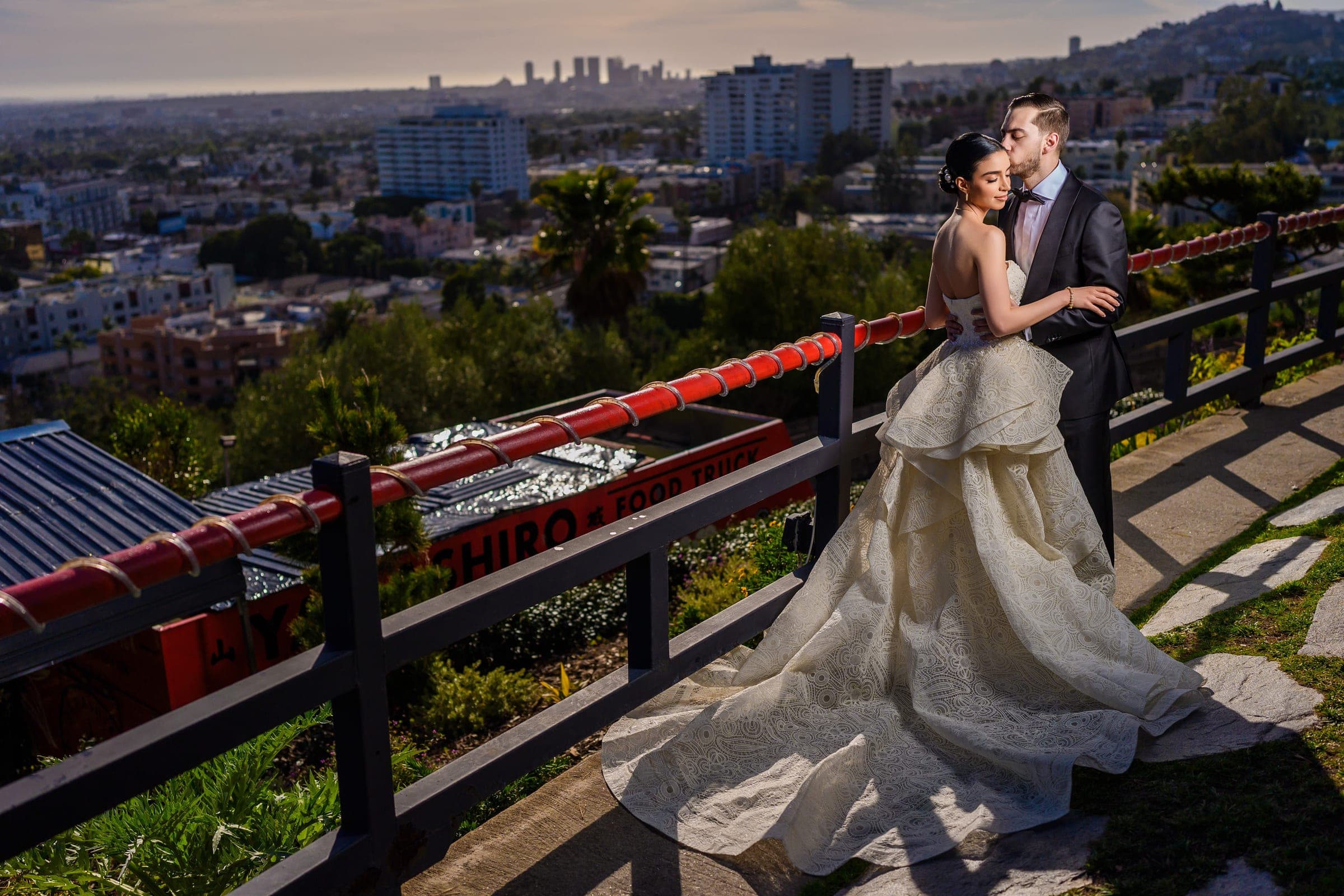 The groom kisses the bride at a hilltop railing, the city grid and distant towers spread below them.
