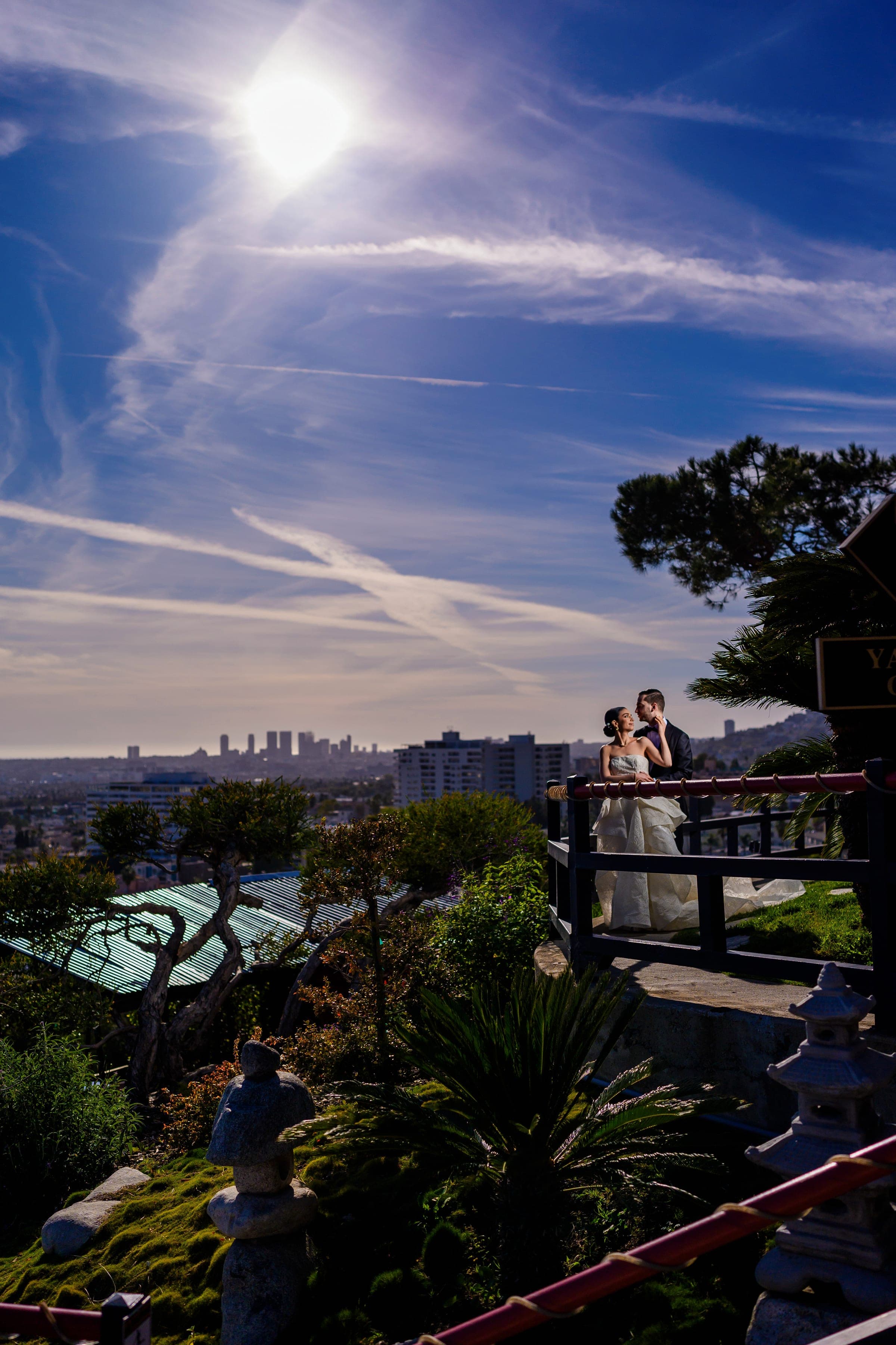 The couple embraces at a hilltop garden railing beneath a bright sun, the Los Angeles skyline behind them.