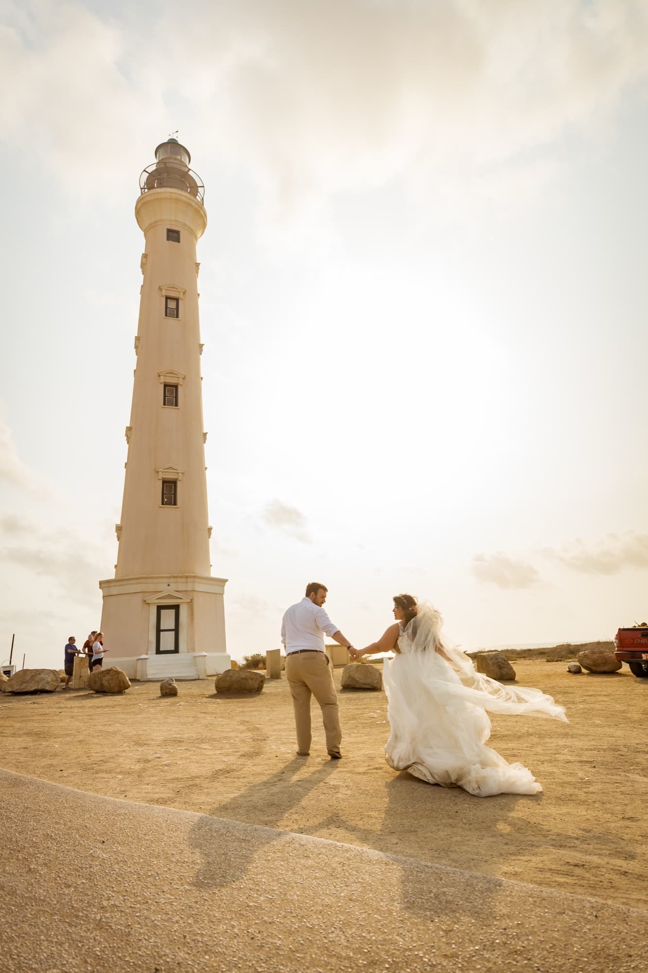 Nikole and Nick holding hands at the base of the white California Lighthouse in Aruba, her train sweeping across the sand