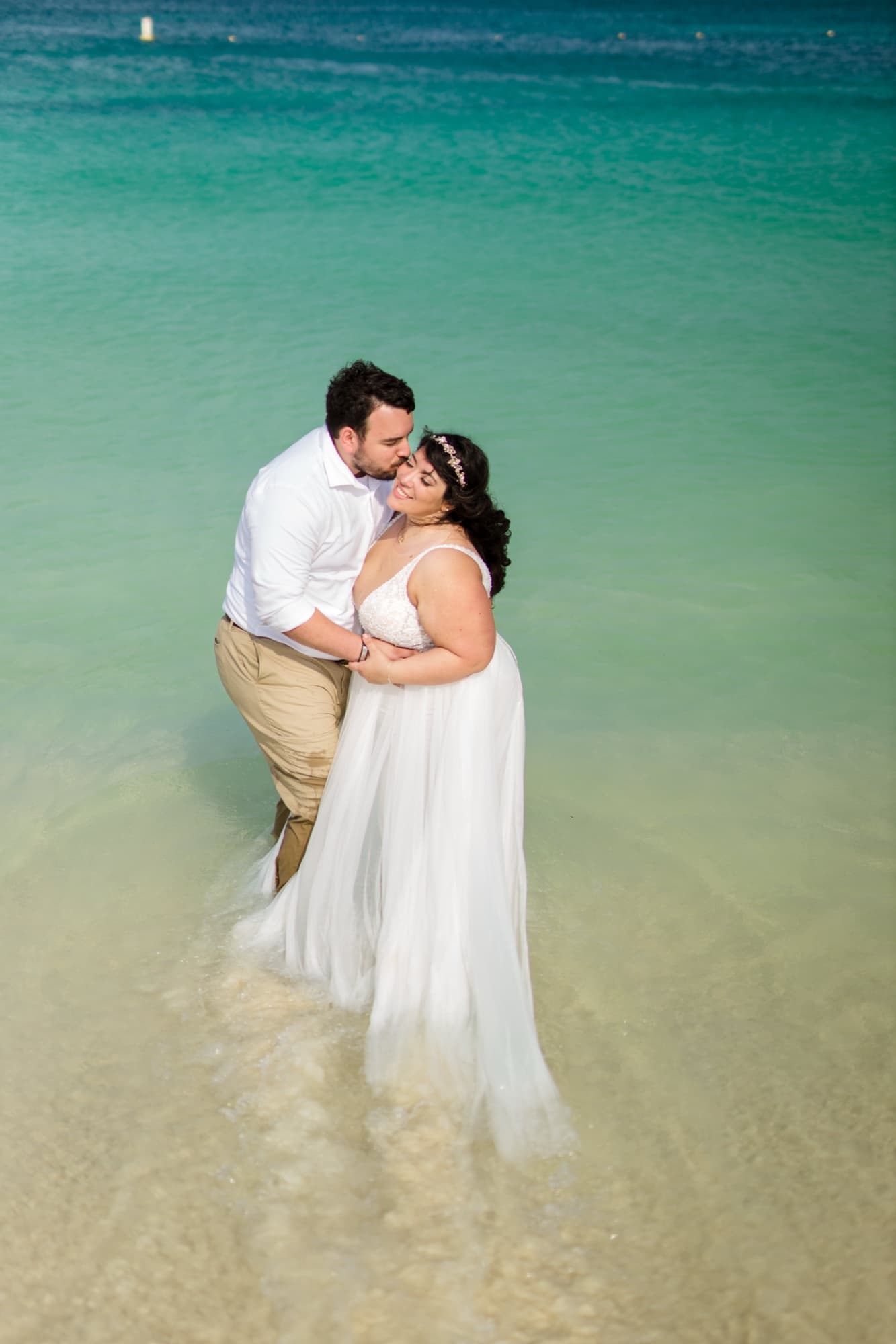 Nikole and Nick standing in the clear turquoise shallows of Eagle Beach in Aruba, her gown floating on the water
