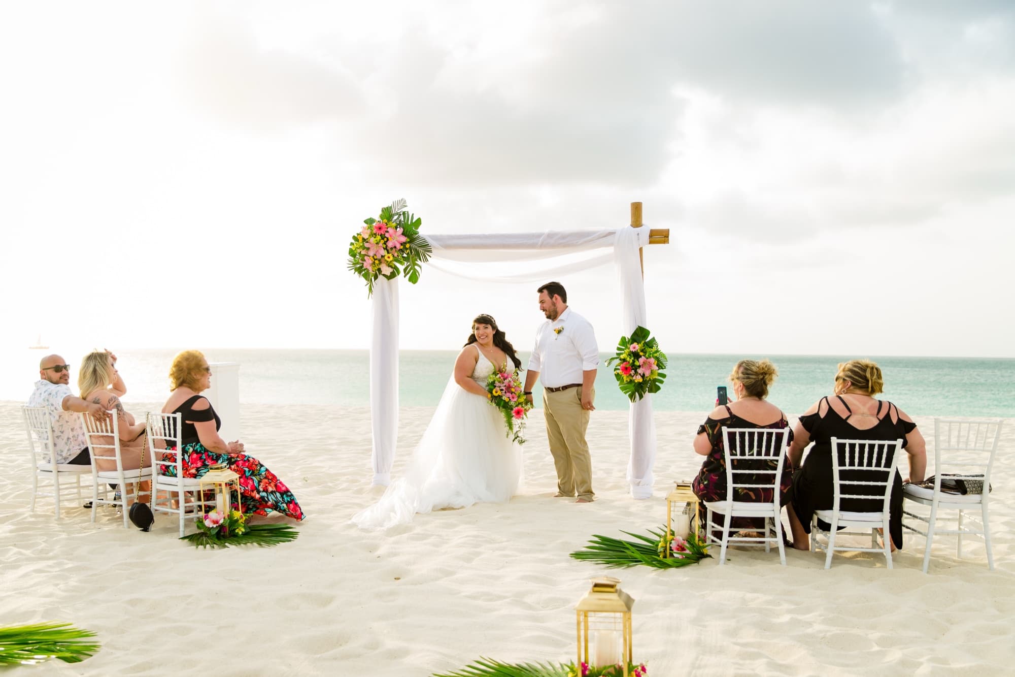 Barefoot beach ceremony under a white gauze arch with tropical flowers on Eagle Beach in Aruba, guests seated in the sand