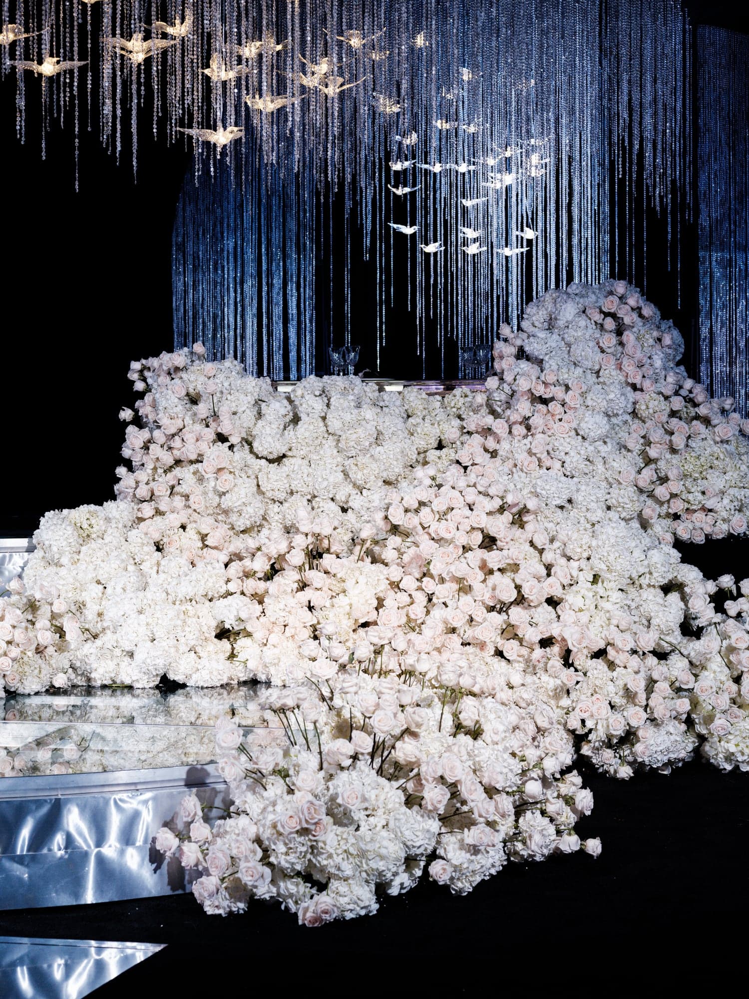 A tall white wedding cake displayed beneath crystal lighting in the Beverly Hilton ballroom