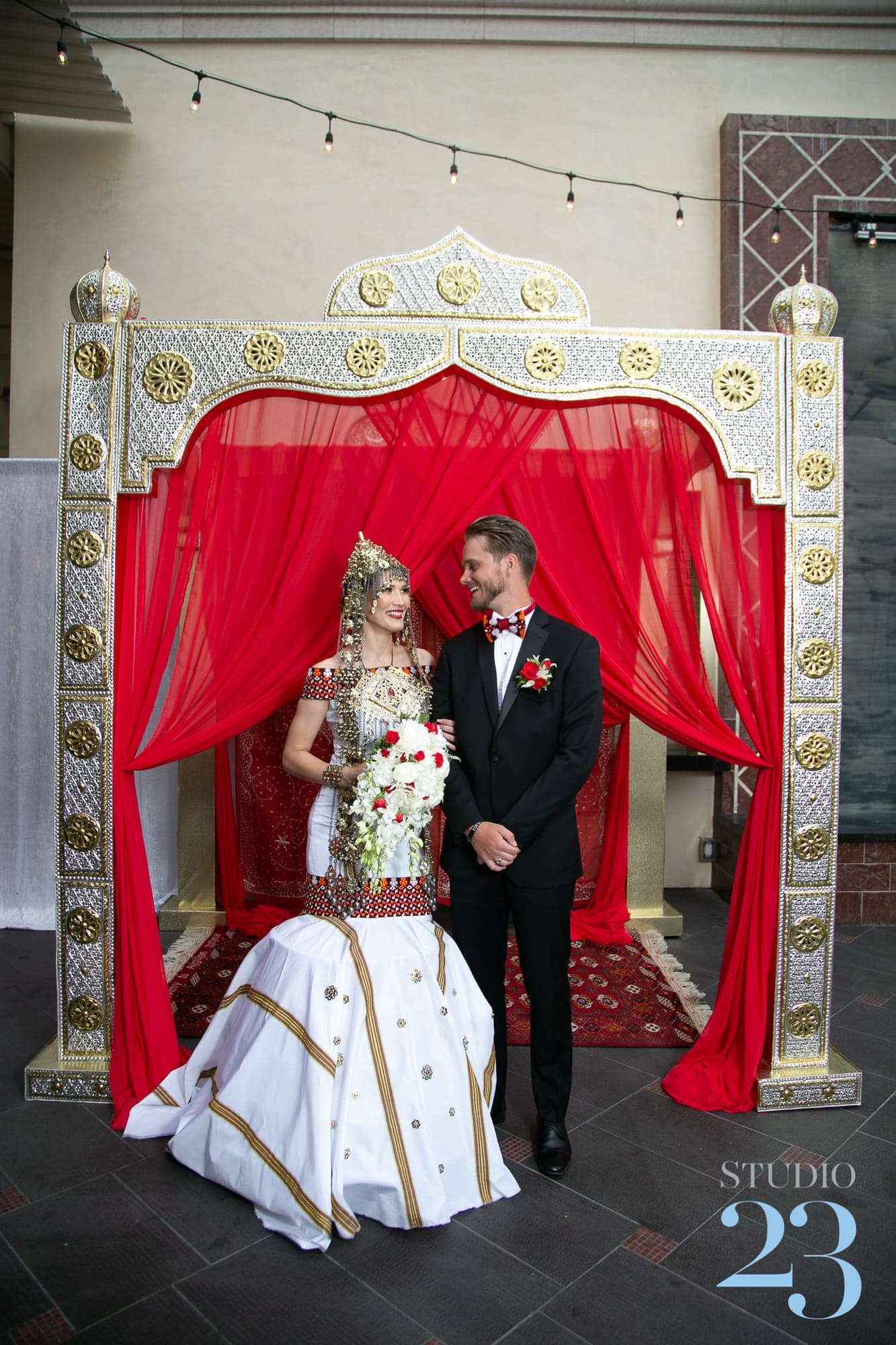 Aygul and Cameron beneath a red ceremonial wedding backdrop