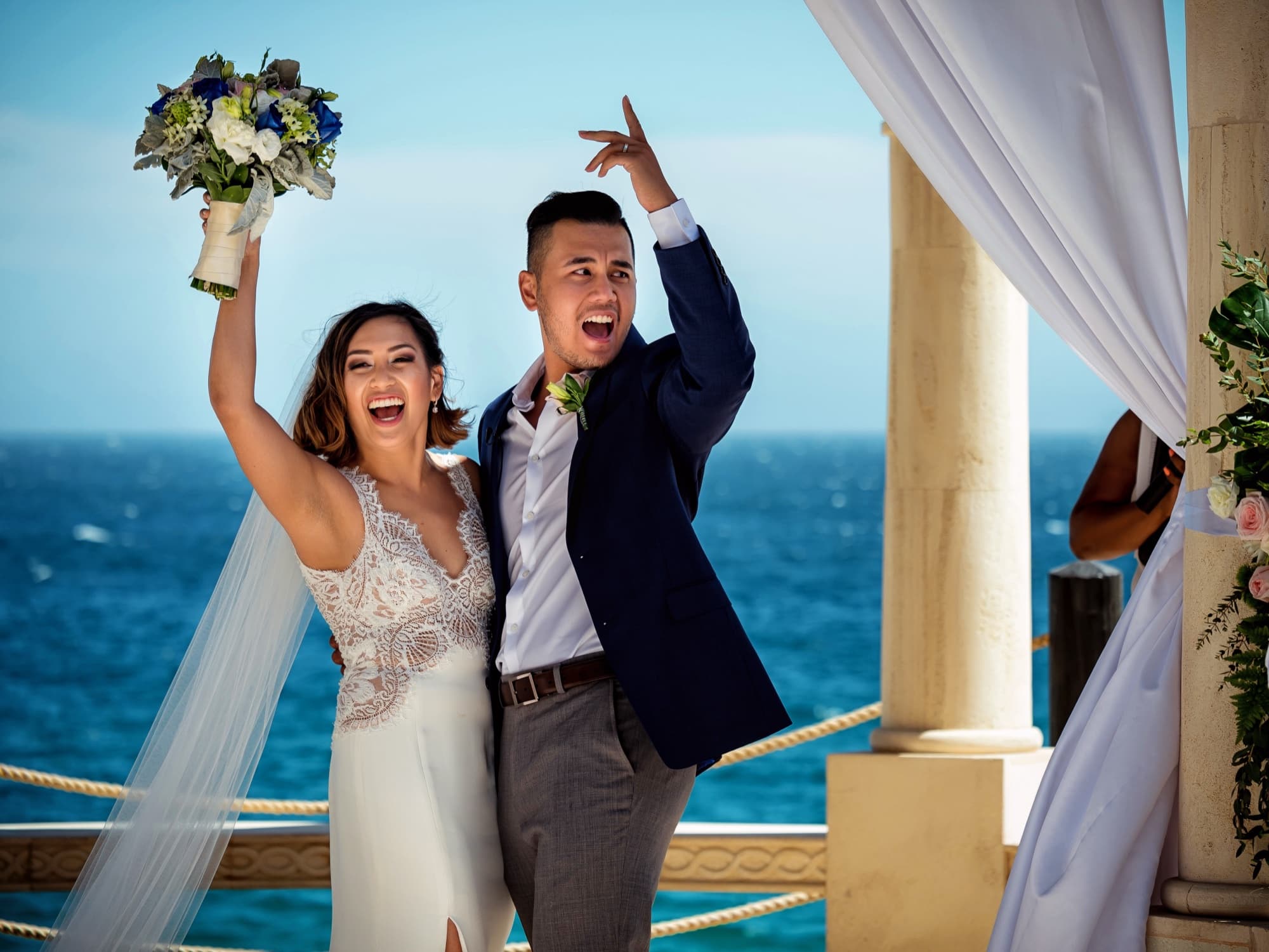 Kirsten and Jolan cheering with her bouquet raised at the ocean-view pergola after their Cabo ceremony