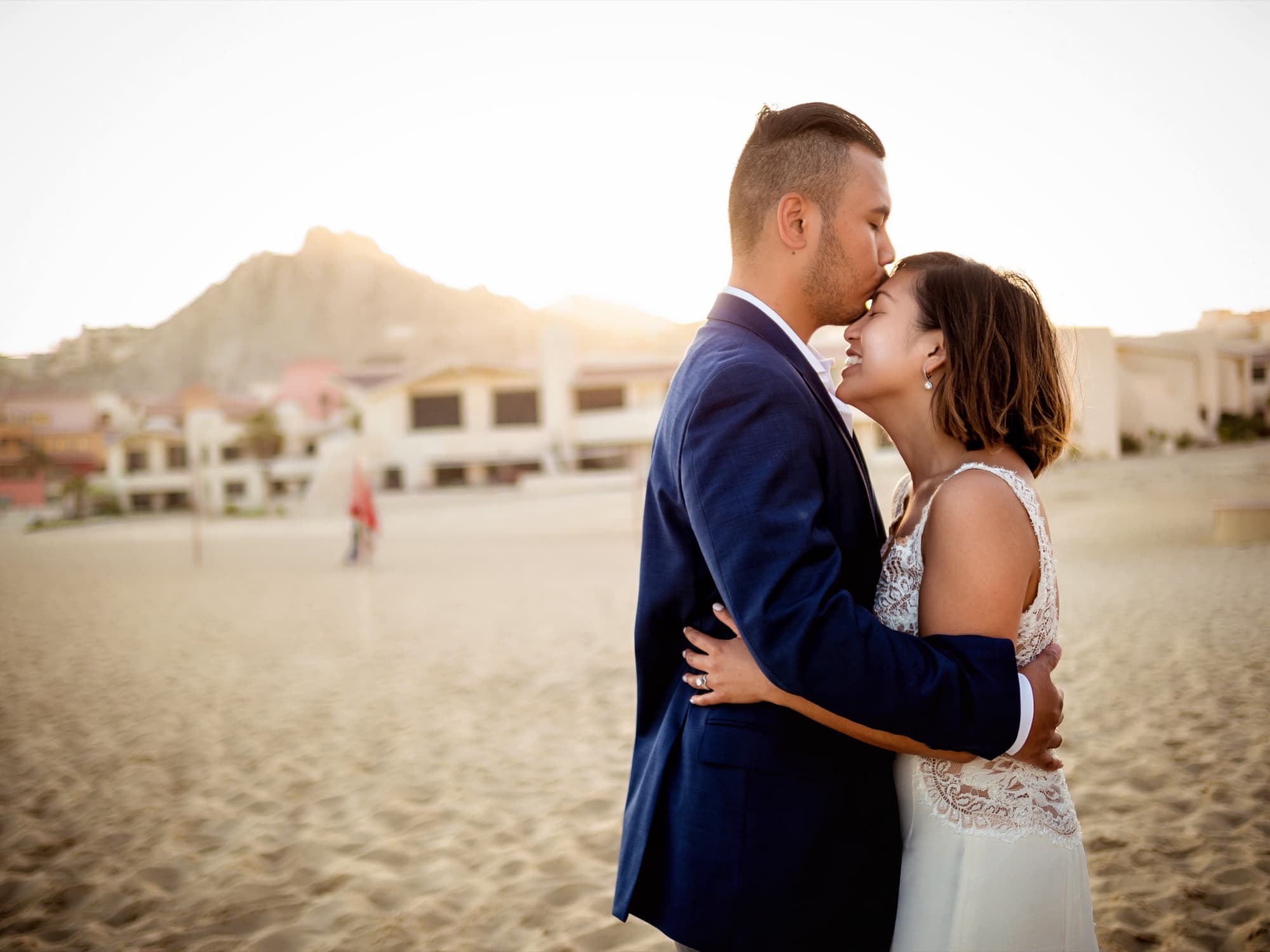 Jolan kissing Kirsten's forehead on the beach at sunset with the Cabo mountains glowing behind them