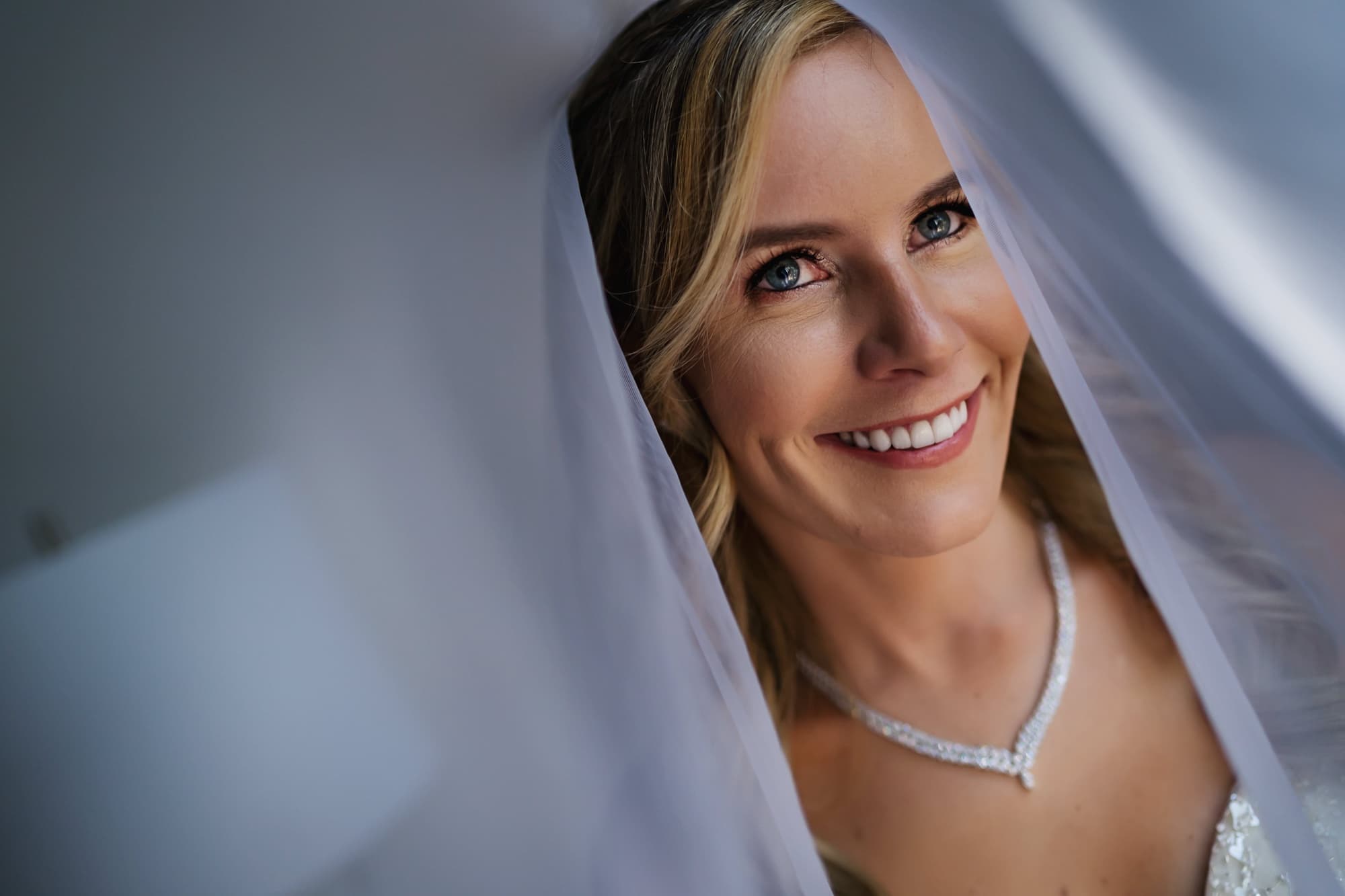 Monica smiling beneath her veil with a diamond necklace, soft window light wrapping her face