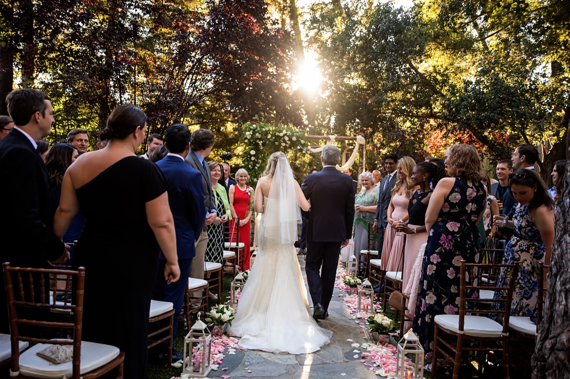 Monica walking the petal-lined aisle on her father's arm as evening sun bursts through the sycamores at Calamigos Ranch