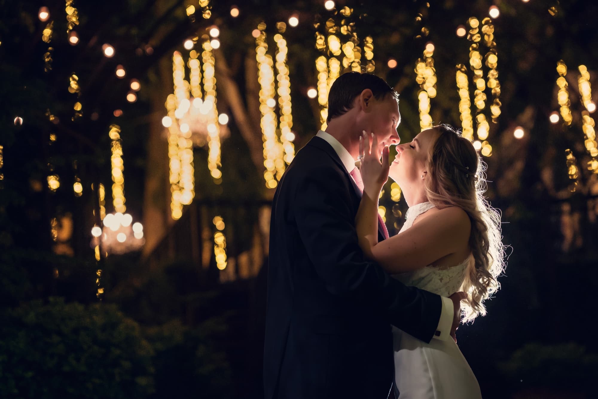 Monica and Colin kissing at night beneath Calamigos Ranch's hanging twinkle lights and chandeliers