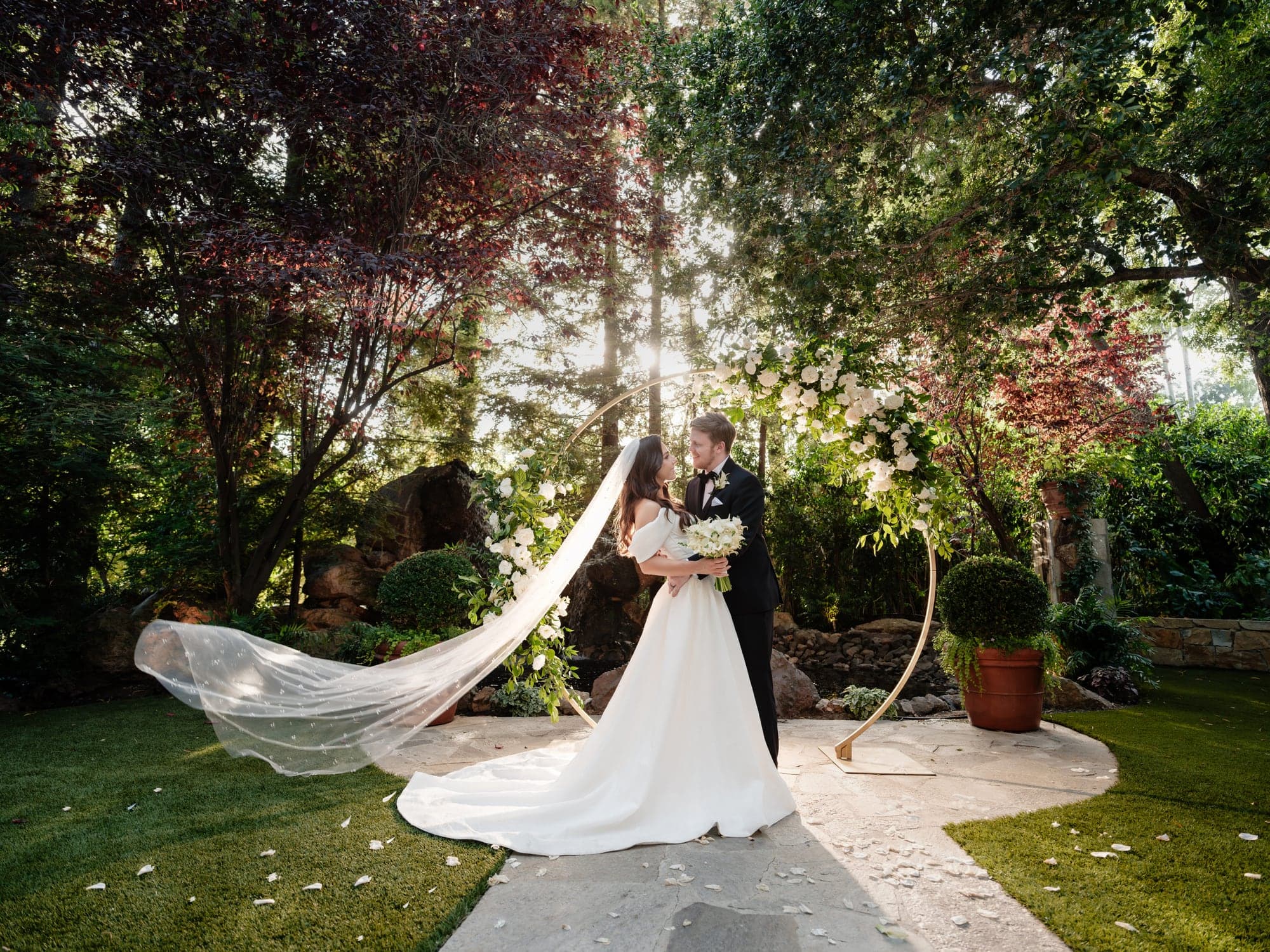 The couple walking through warm garden light with the bride's veil trailing behind