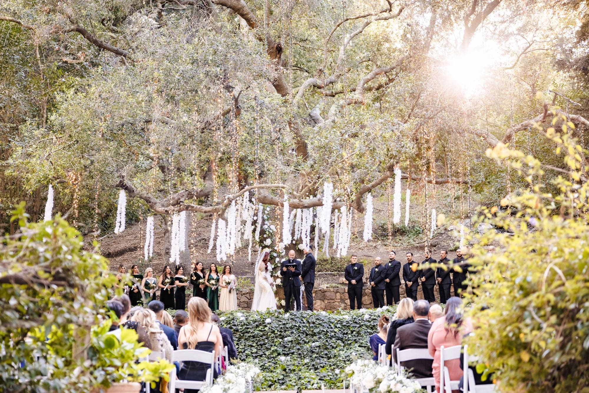 Gina walking toward the Calamigos Ranch ceremony through warm light beneath the trees