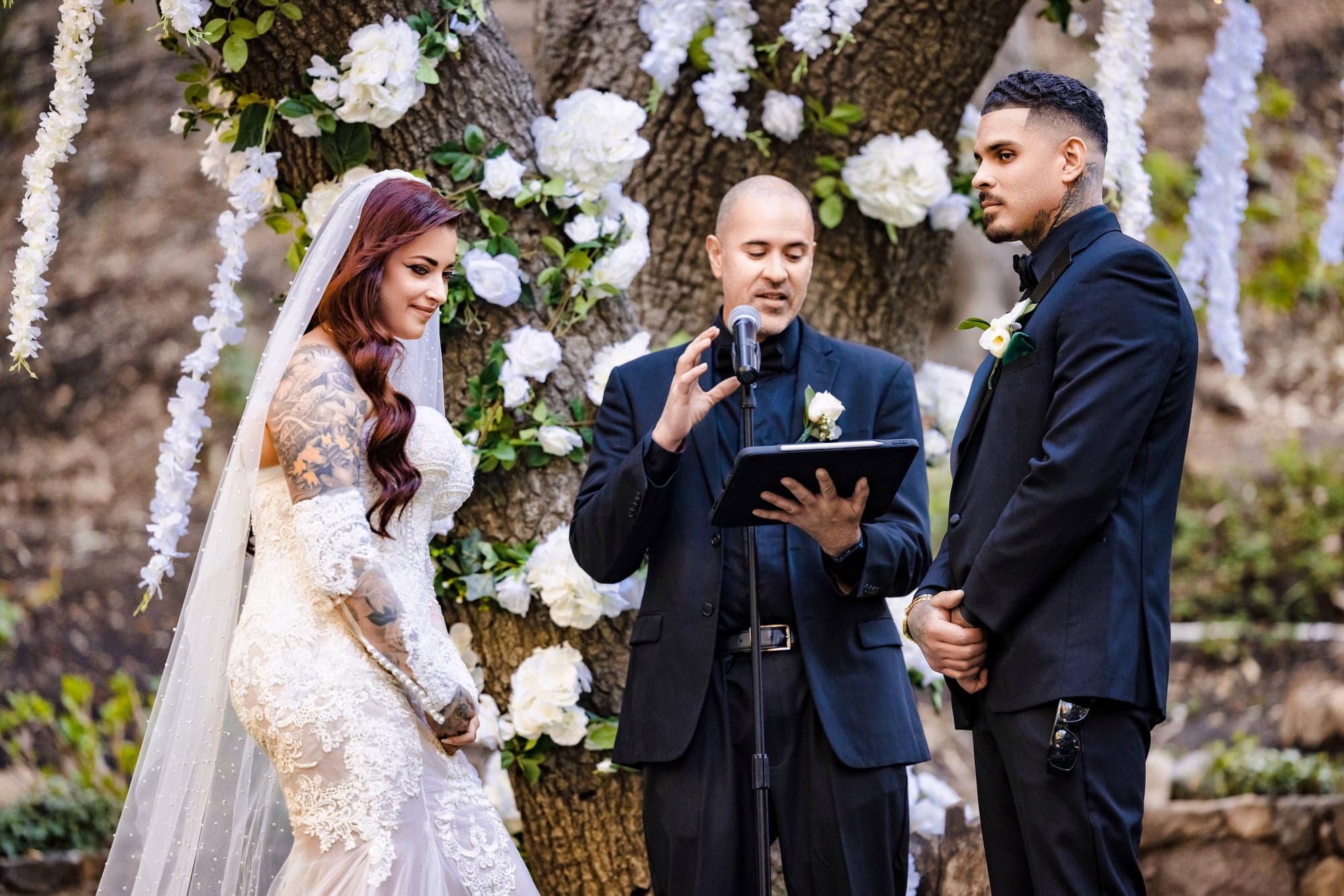 Gina and Alonso exchanging vows beneath a floral installation in the Calamigos oak grove
