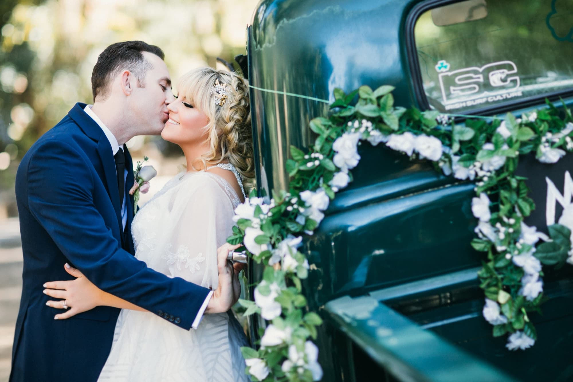 Groom kissing the bride beside a vintage green pickup truck dressed with white floral garland at Calamigos Ranch