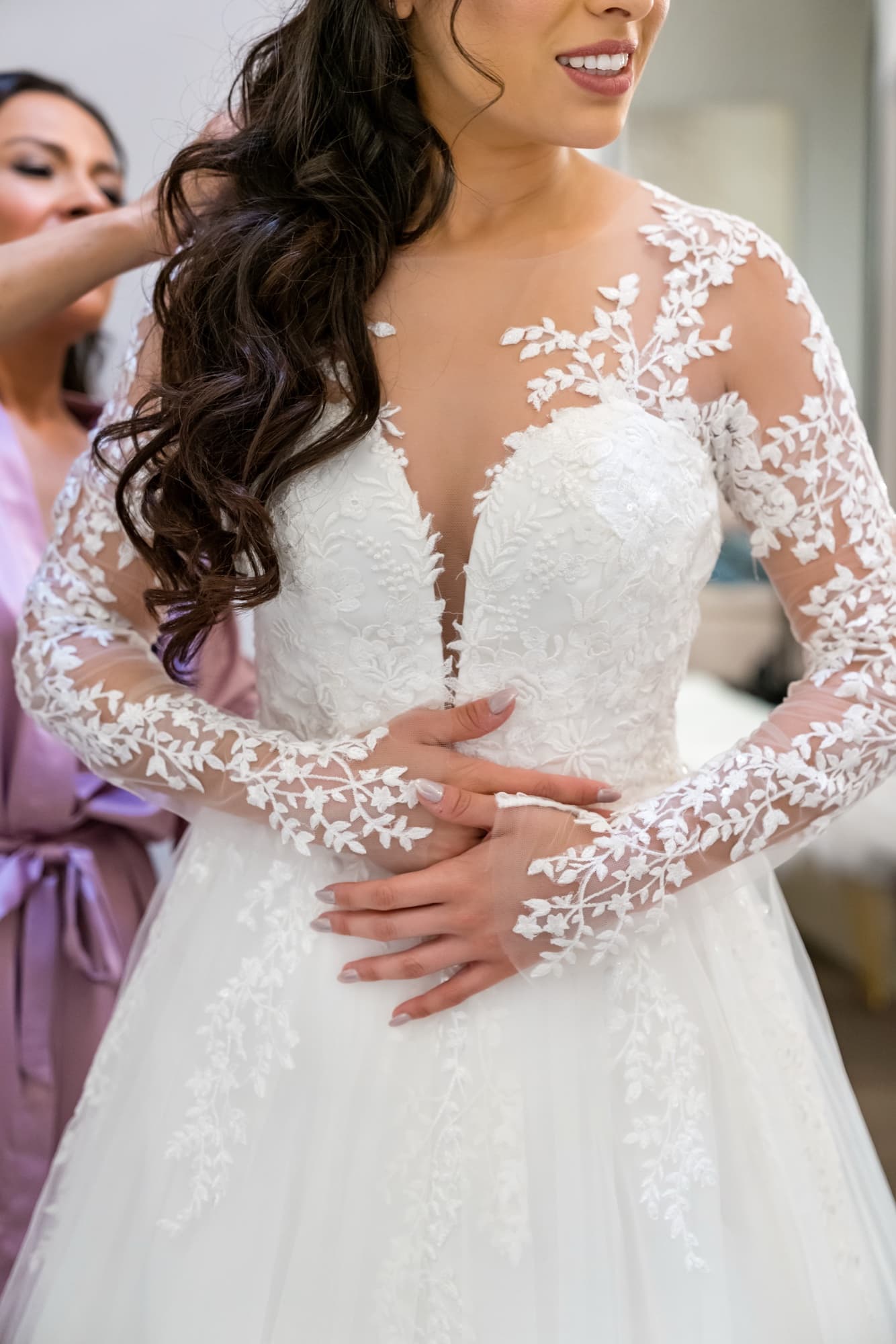 Close view of Tania's illustrated-lace long-sleeve ballgown as her bridesmaid adjusts it before the ceremony