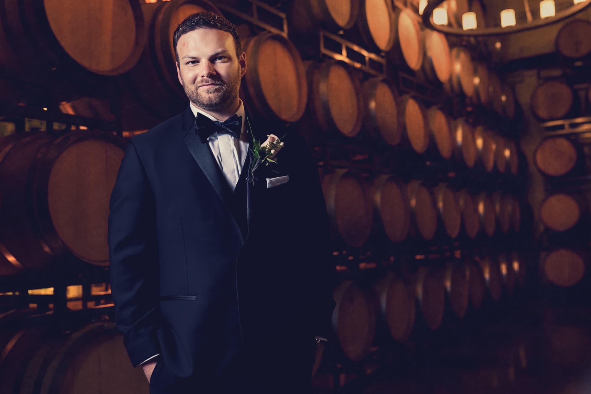 Cole in a navy tuxedo standing before the glowing wall of stacked oak barrels in the Callaway barrel room