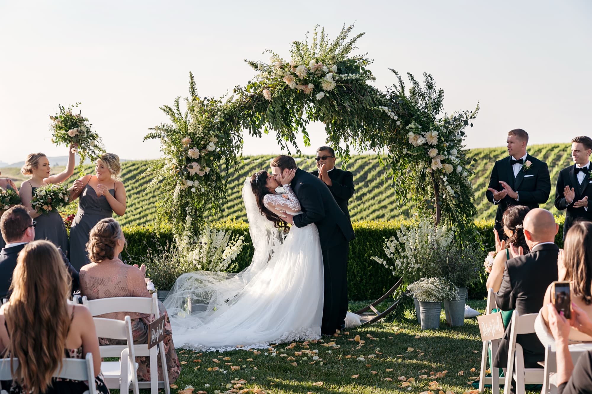 Tania and Cole kissing under the greenery ceremony arch with the Temecula vineyard rows behind them