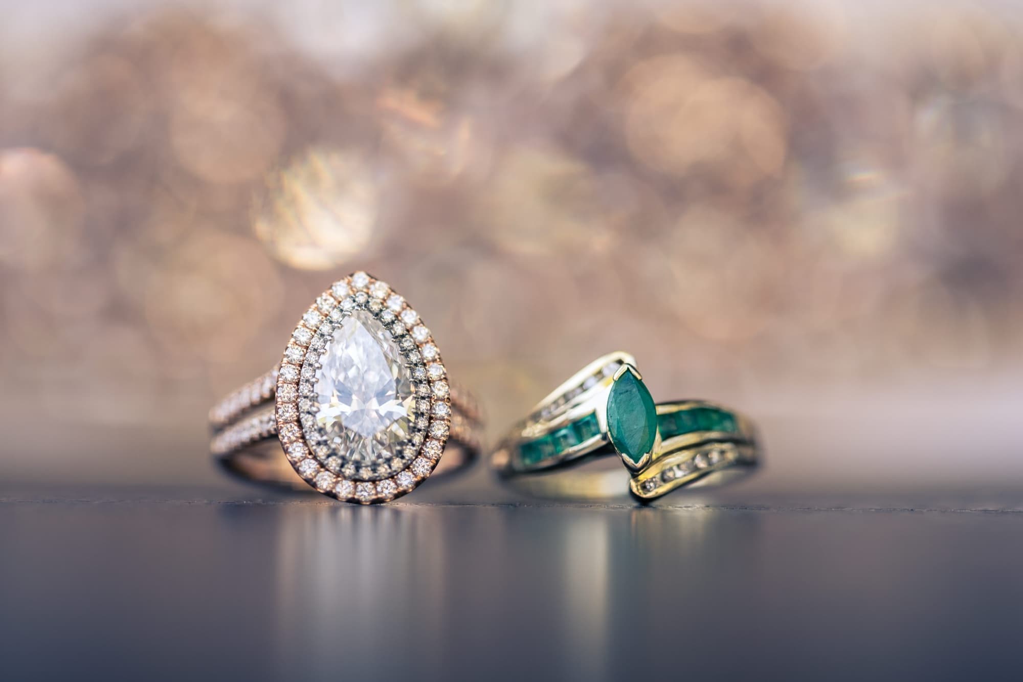 Double-halo pear diamond engagement ring beside a gold band set with emeralds on a reflective surface at Carondelet House