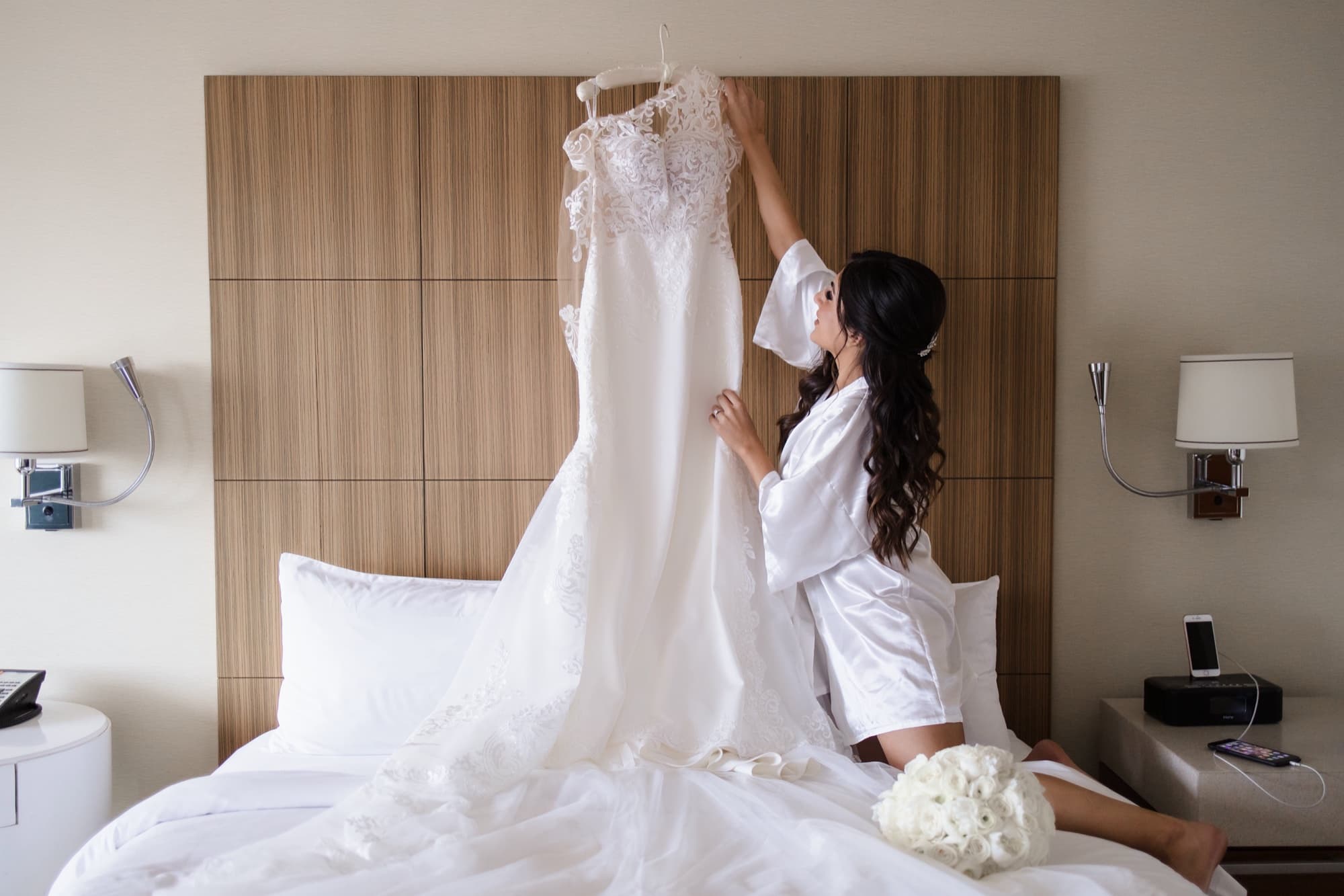 Hela in her white robe reaching up for the lace wedding gown hanging above the hotel bed
