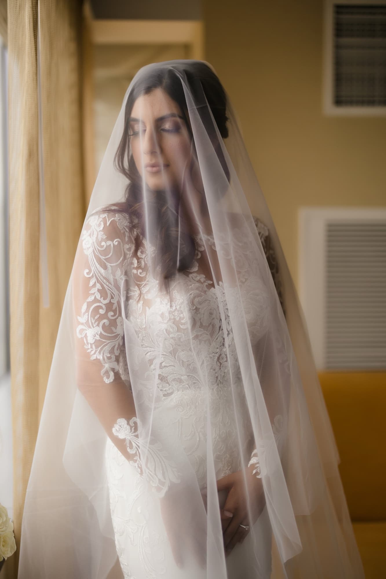 Hela beneath her cathedral veil in the window light, eyes closed in a quiet moment before the ceremony
