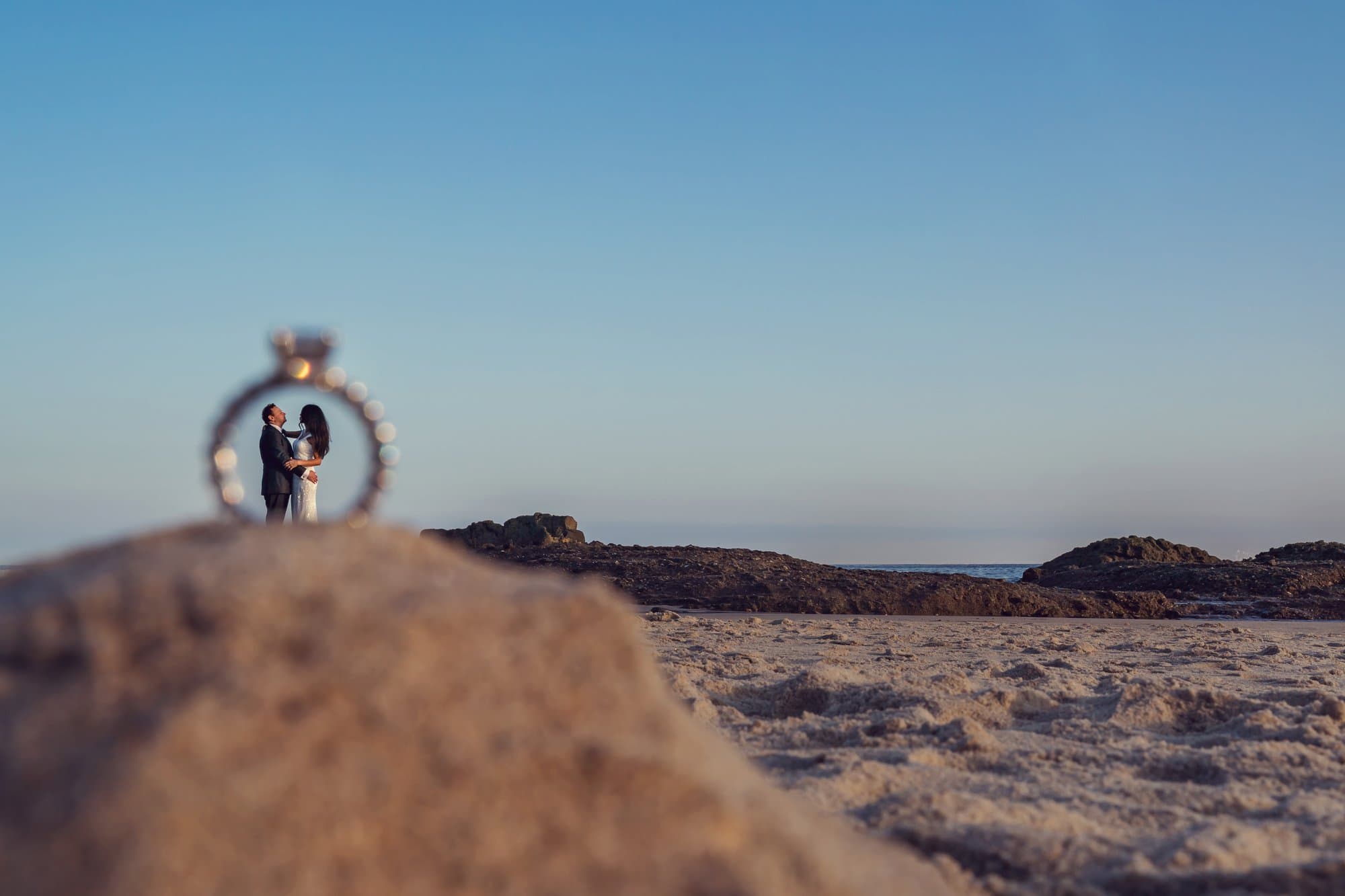 An engagement ring balanced on a beach rock in the foreground, with the couple embracing framed inside the band