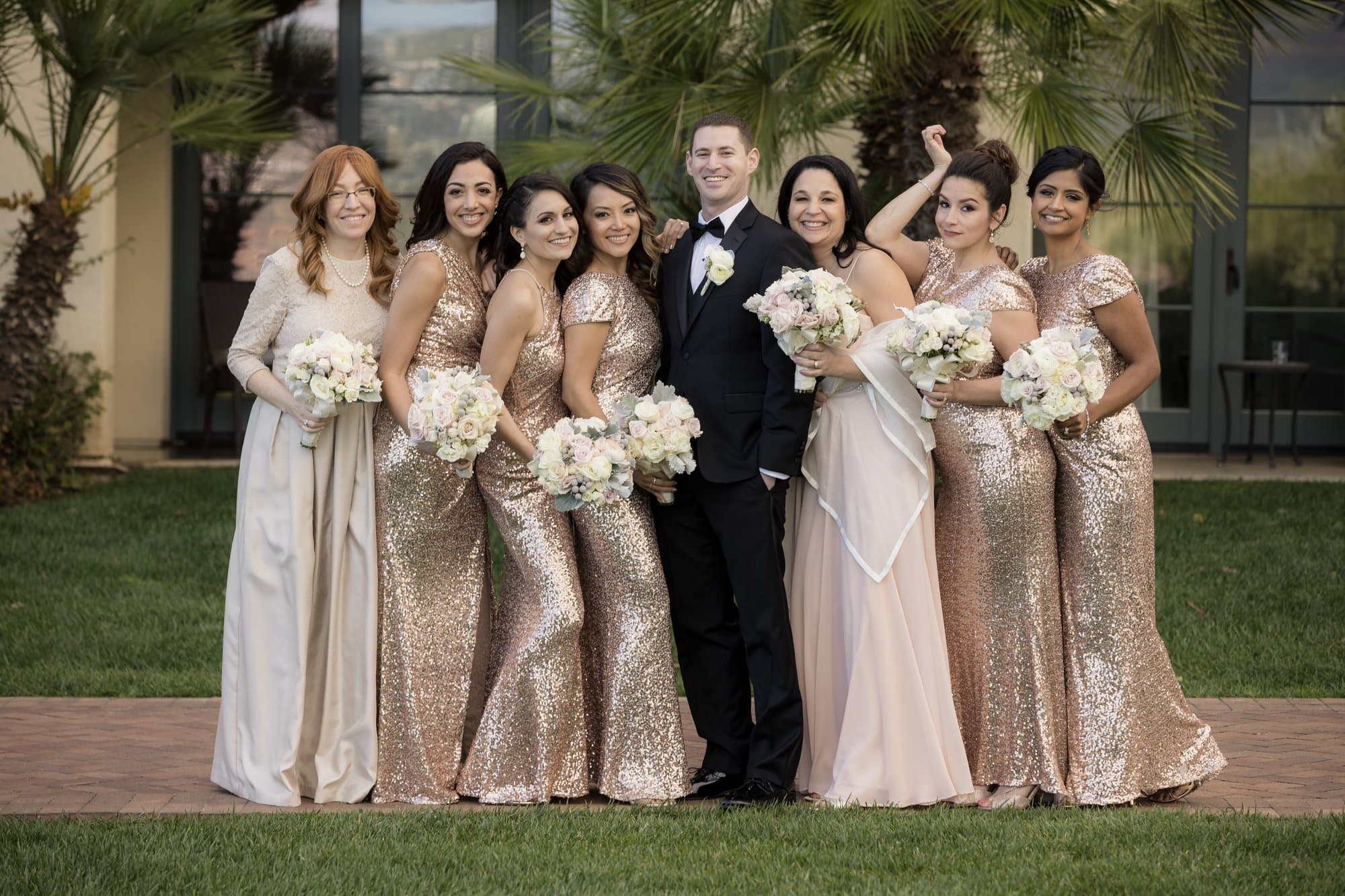 A groom surrounded by bridesmaids in gold sequined gowns striking playful poses on a resort lawn