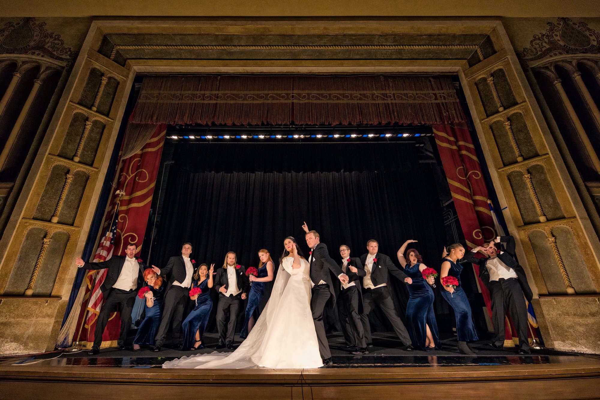 A wedding party striking playful poses with the couple at center stage in a historic theater
