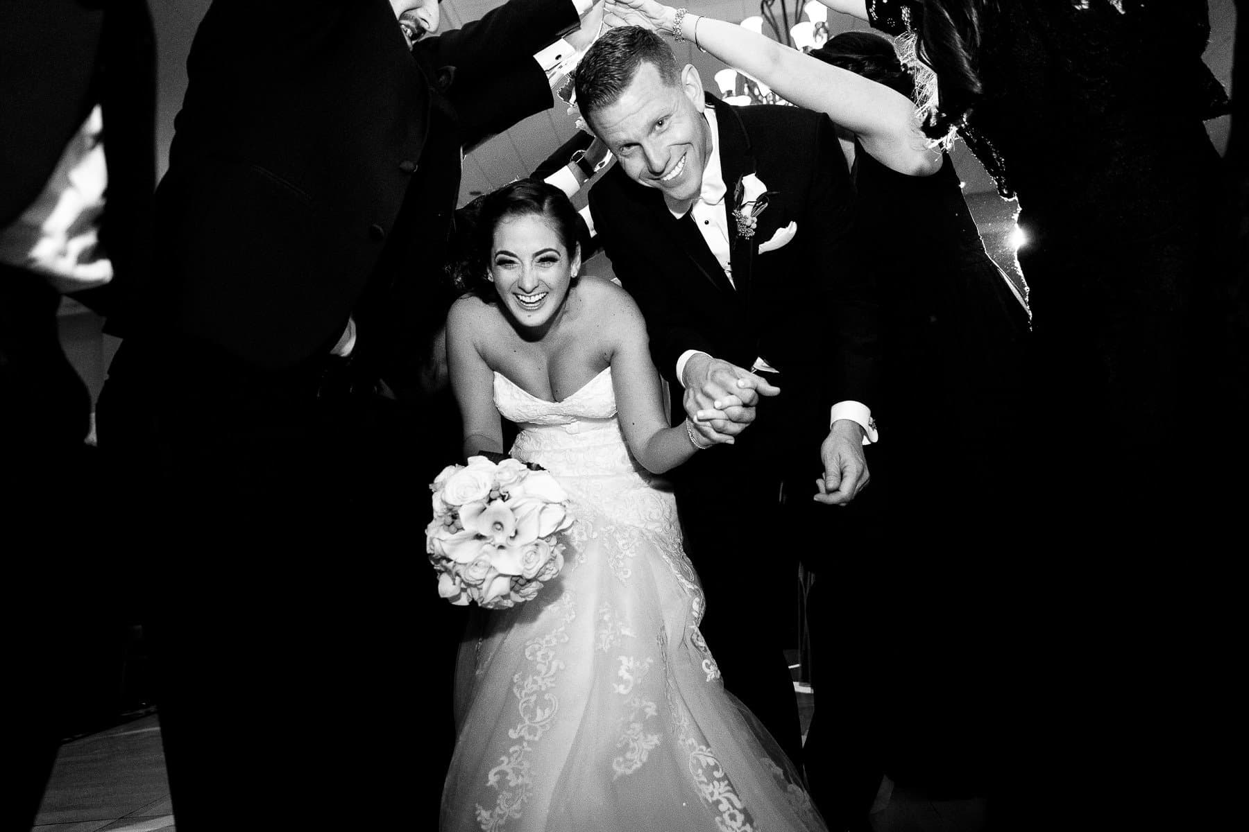 Black and white photograph of a bride and groom grinning as they duck through a tunnel of guests' arms