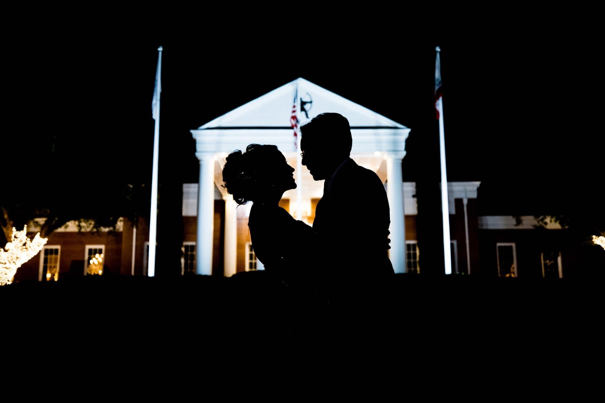 A bride and groom kissing in silhouette at night before the illuminated columns of a country club