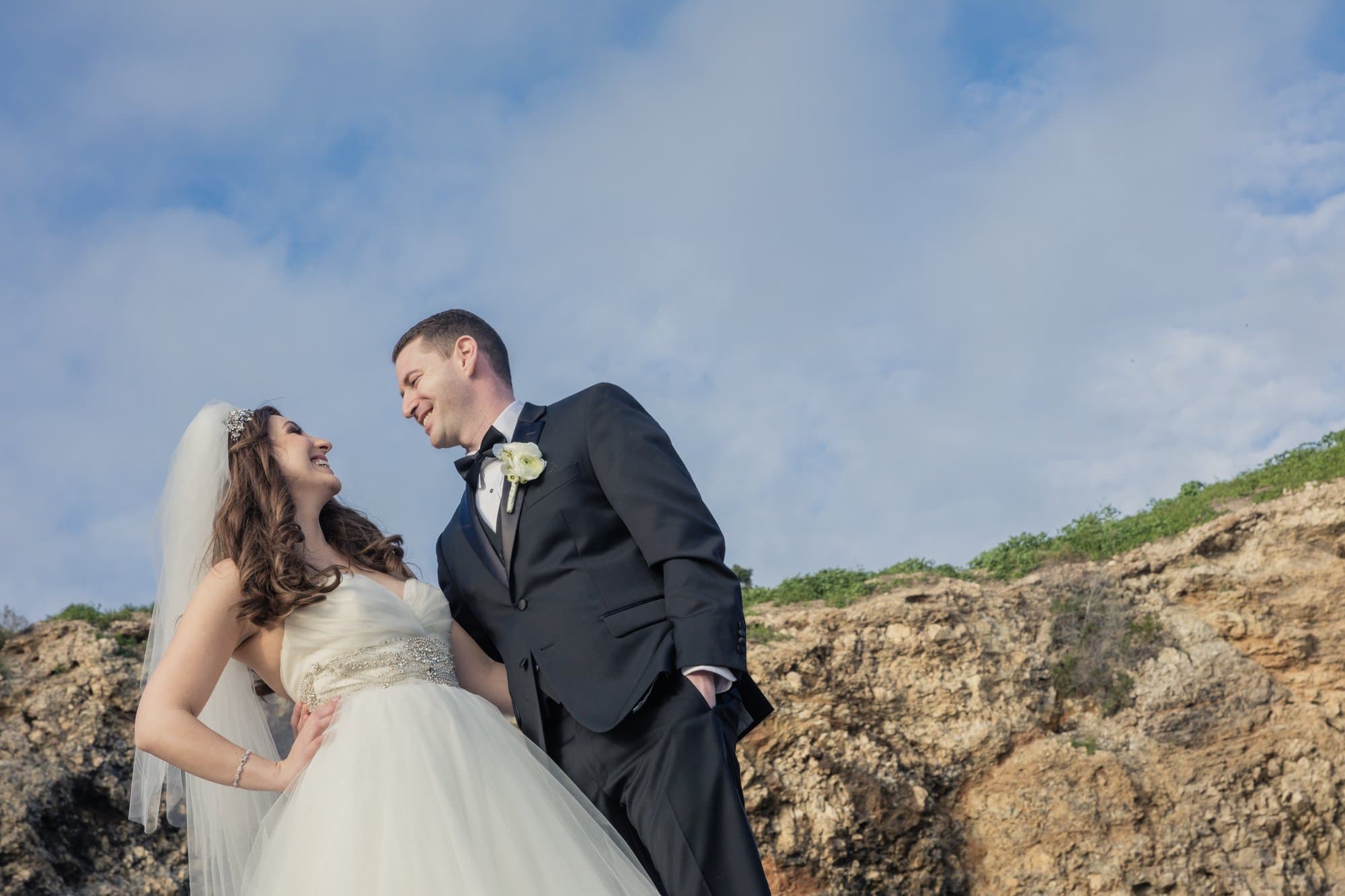 A bride and groom laughing together on coastal rocks under a bright blue sky