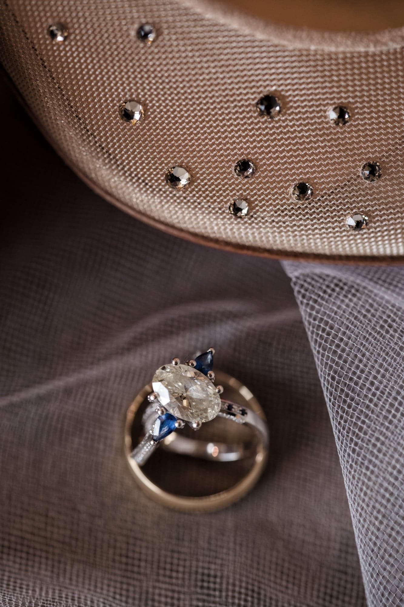 Erin's oval engagement ring with blue side stones and the wedding bands resting on a studded hat brim