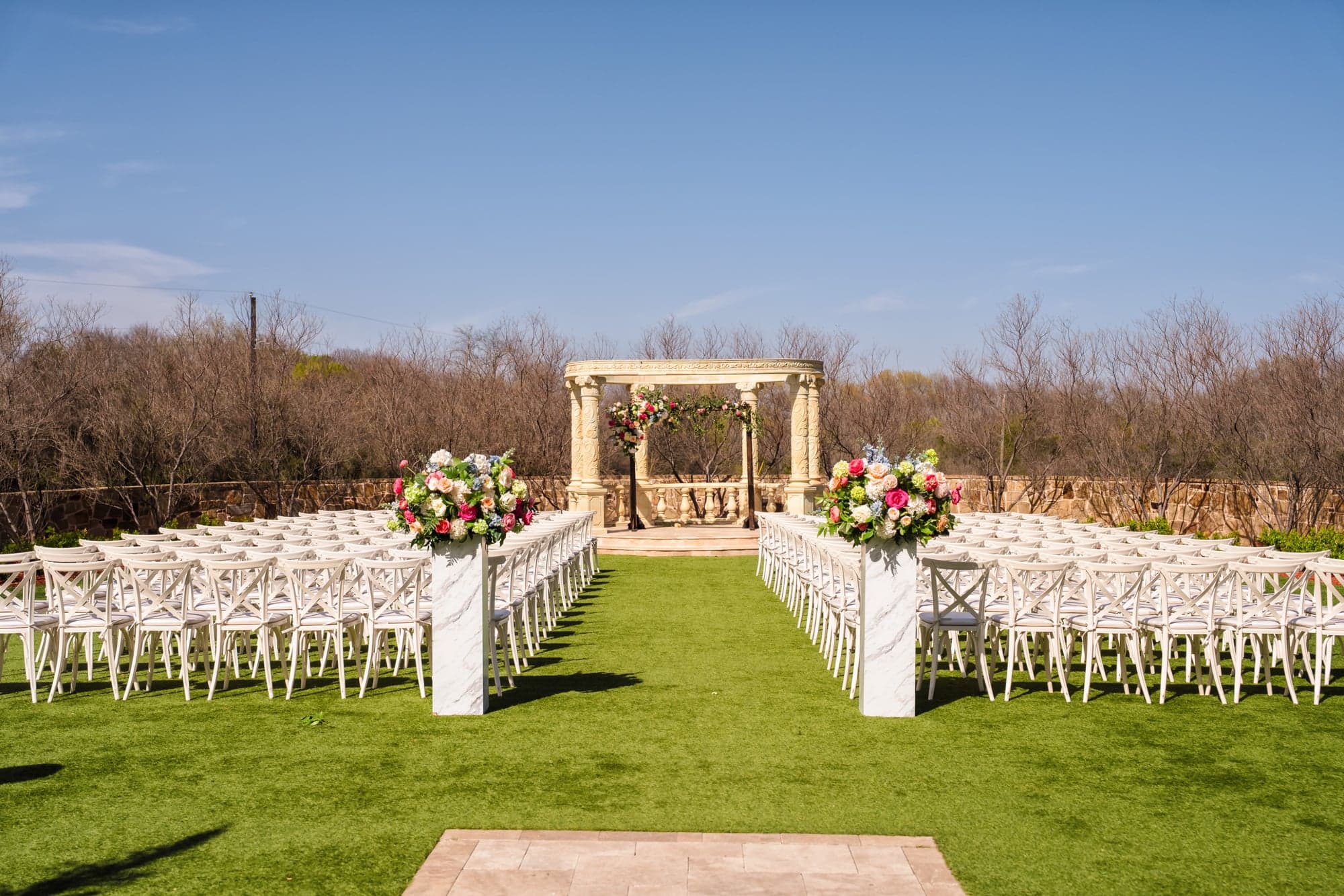 The ceremony lawn at D'Vine Grace Vineyard set with white cross-back chairs facing the floral-wrapped stone rotunda