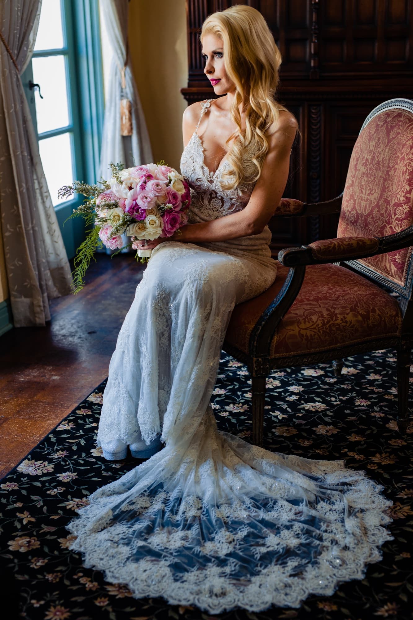 Katie seated in an antique chair by the window at the Ebell, her beaded gown's lace train pooled across the floral carpet
