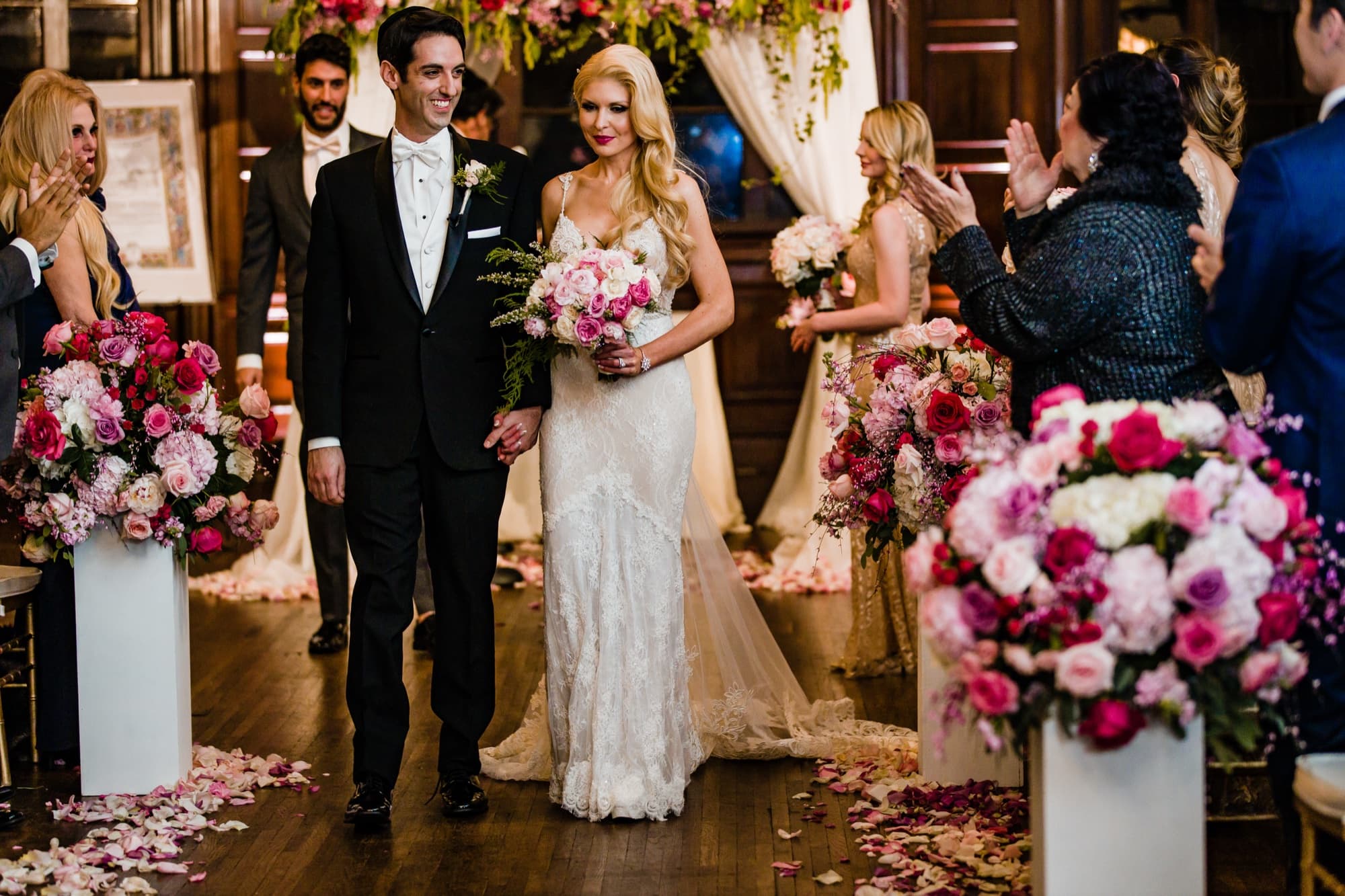 Katie and Blake walking the petal-strewn aisle between towers of pink and fuchsia roses in the grand hall of the Ebell