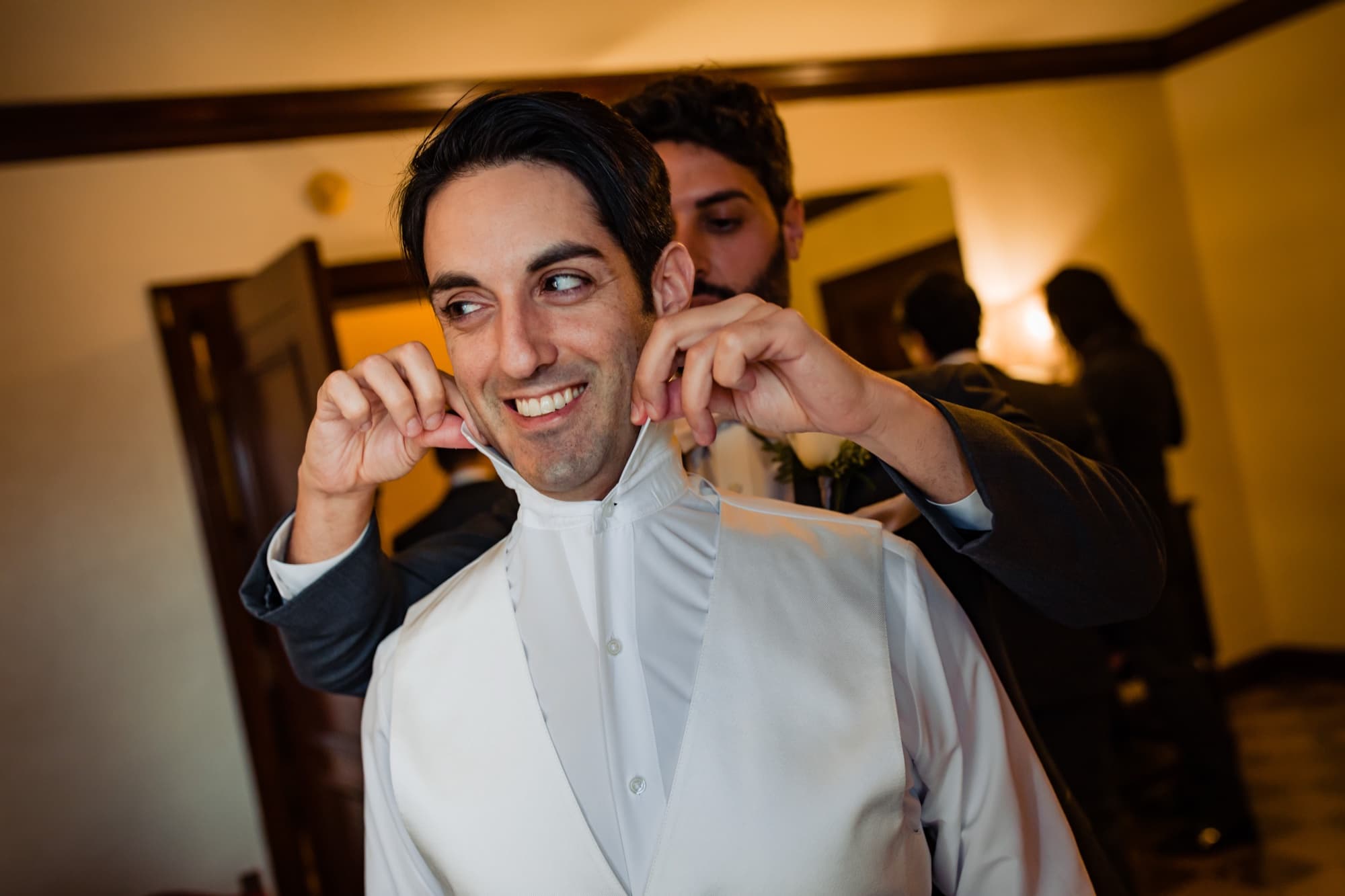 Blake grinning as his collar is adjusted during groom prep in the wood-paneled club rooms
