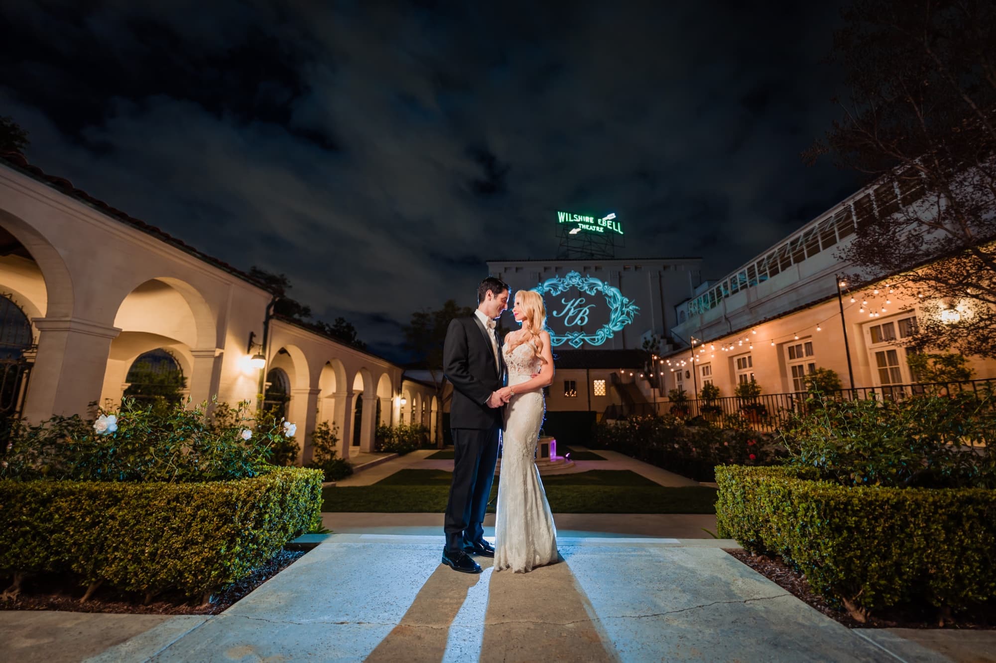 Katie and Blake standing together in the courtyard at night beneath the glowing Wilshire Ebell Theatre sign and their projected monogram, photographed by Michael Anthony Photography