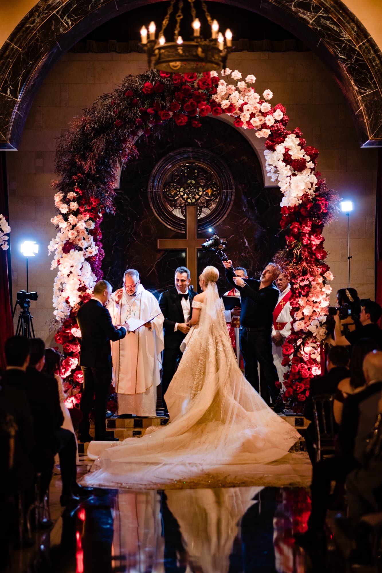 Rhonda and Christopher at the flower-covered ceremony arch with a cross at the Fairmont Grand Del Mar, her train reflected on the polished floor