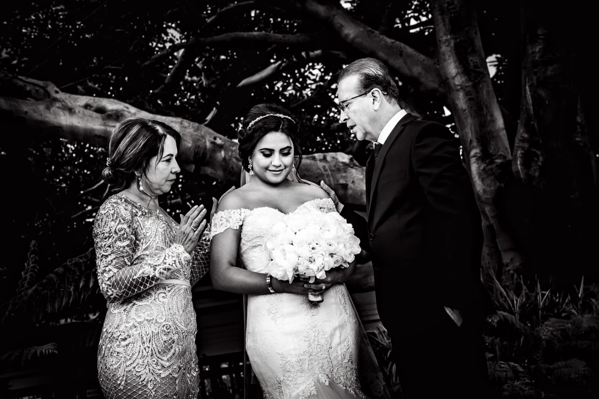 Black and white photograph of Kyrene receiving a blessing from her mother and father beneath the trees before her ceremony