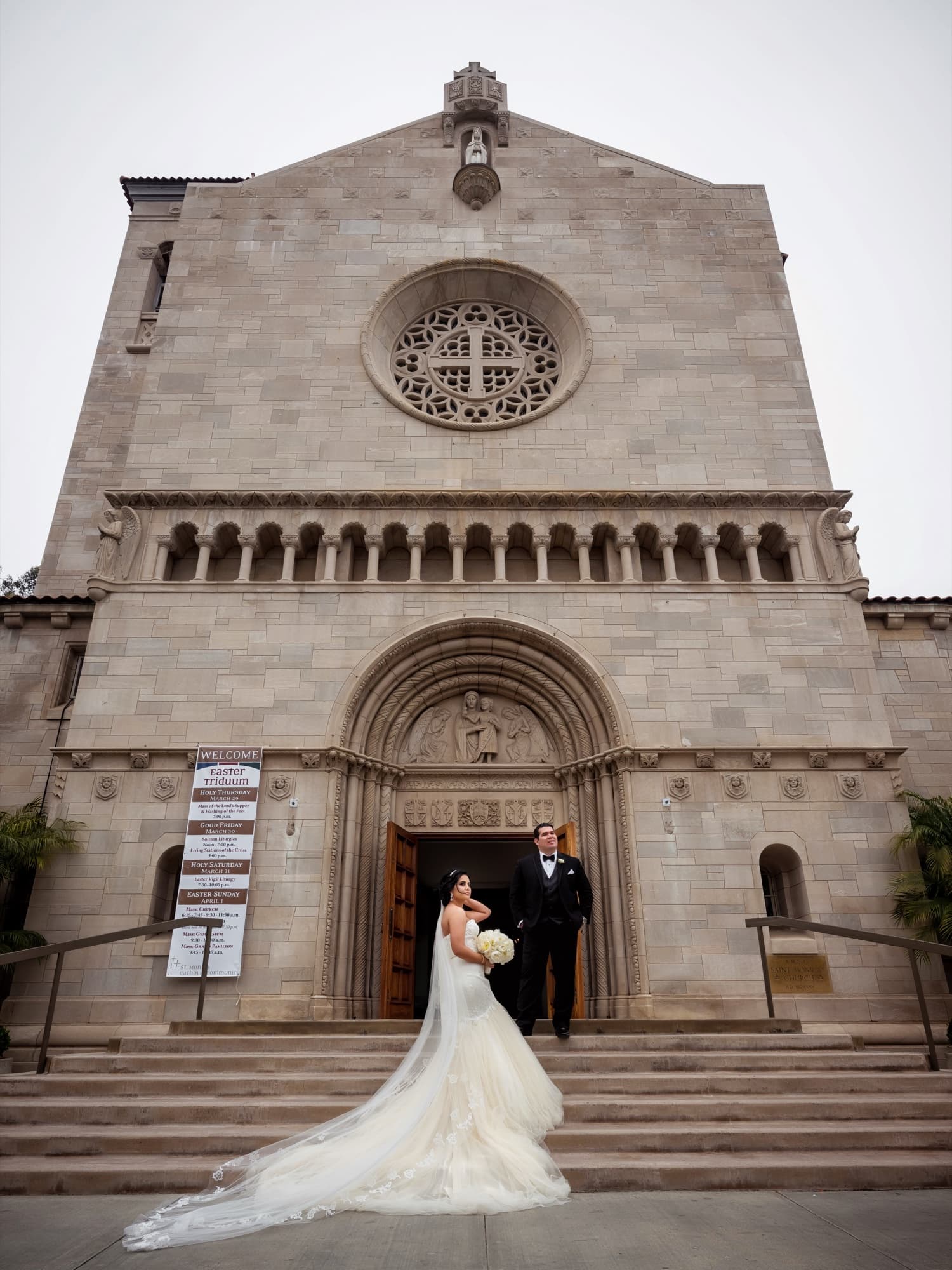 Bride and groom on the front steps of Saint Monica Catholic Church beneath its Romanesque stone facade and rose window