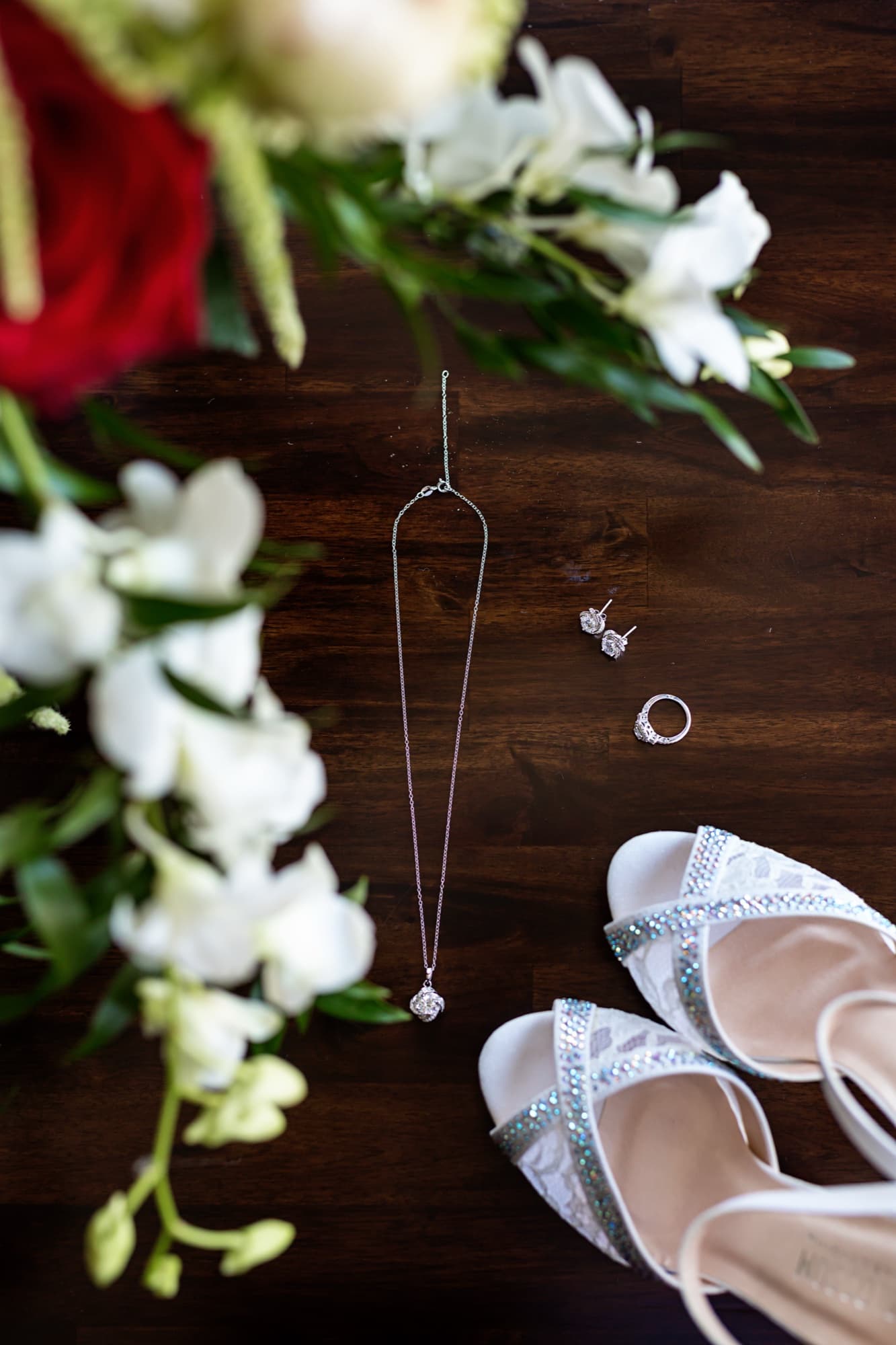 Wedding detail flat lay of crystal-strapped heels, a pendant necklace, rings, and red and white florals on dark wood
