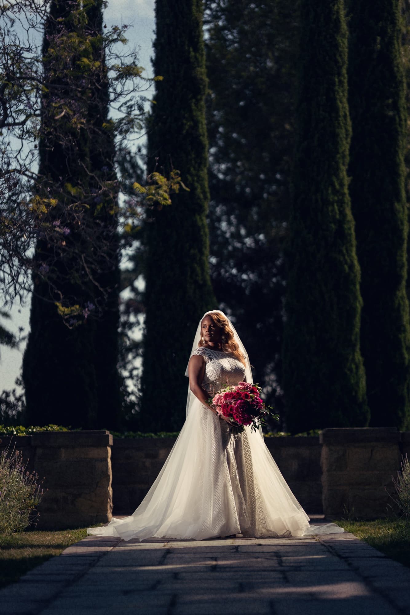 Shaniece standing between towering cypress trees at Greystone Mansion holding a magenta peony bouquet