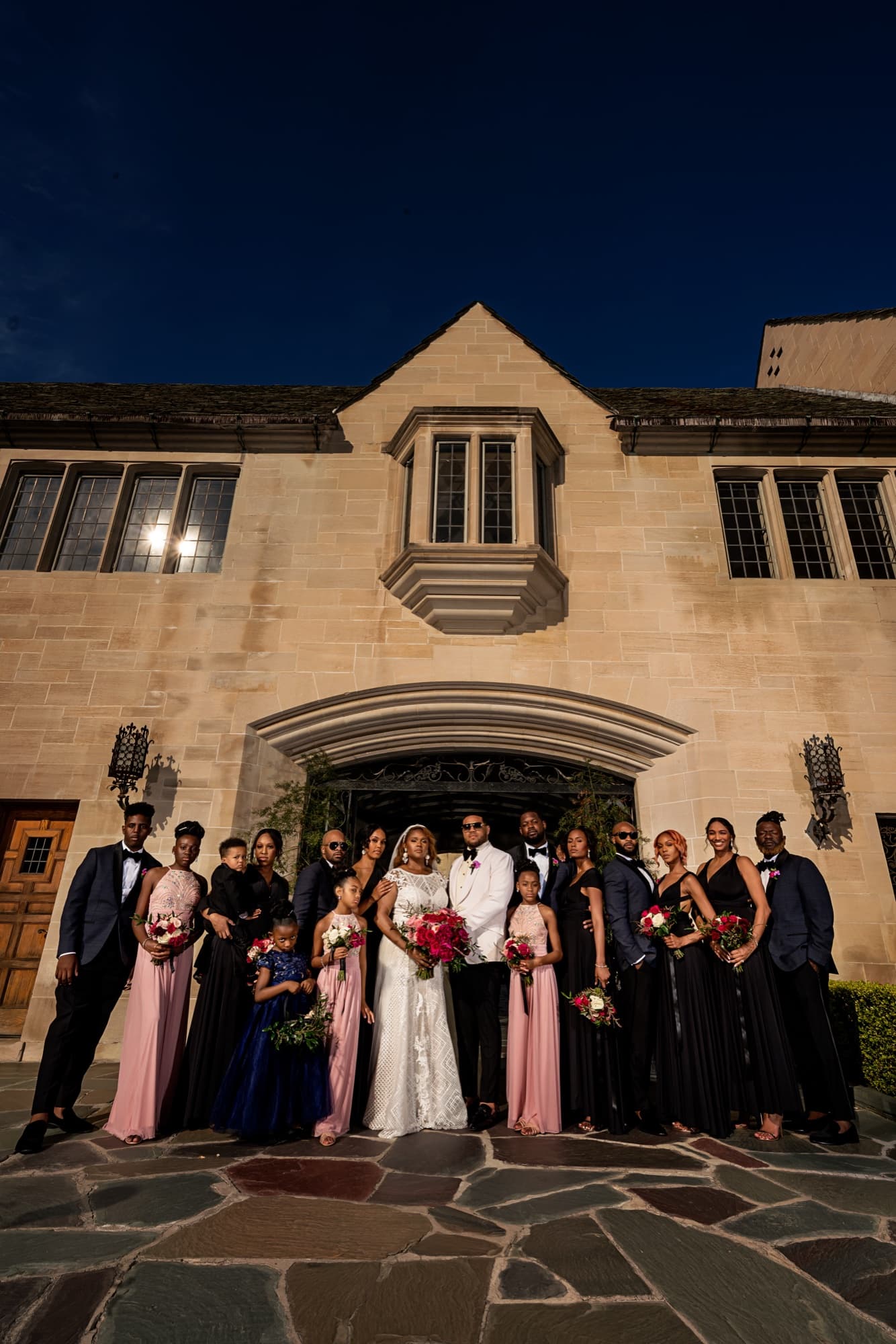 Full wedding party in blush and black gathered at night beneath the lit Tudor facade of Greystone Mansion