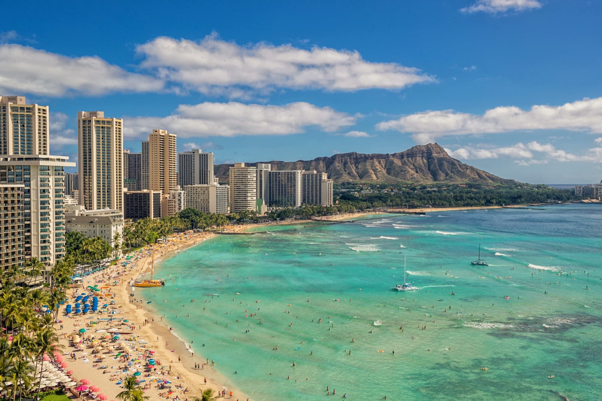 Aerial view of turquoise Waikiki Beach with Diamond Head rising at the far end under a bright blue sky