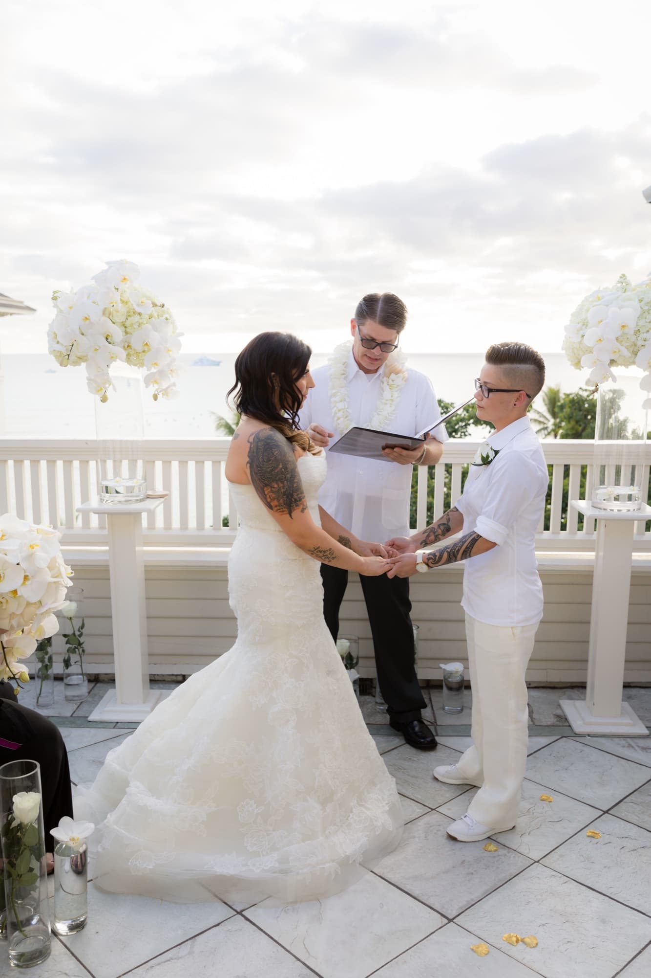 Amanda in a lace gown and Shauna in a white suit holding hands during their ceremony on an oceanfront veranda above Waikiki