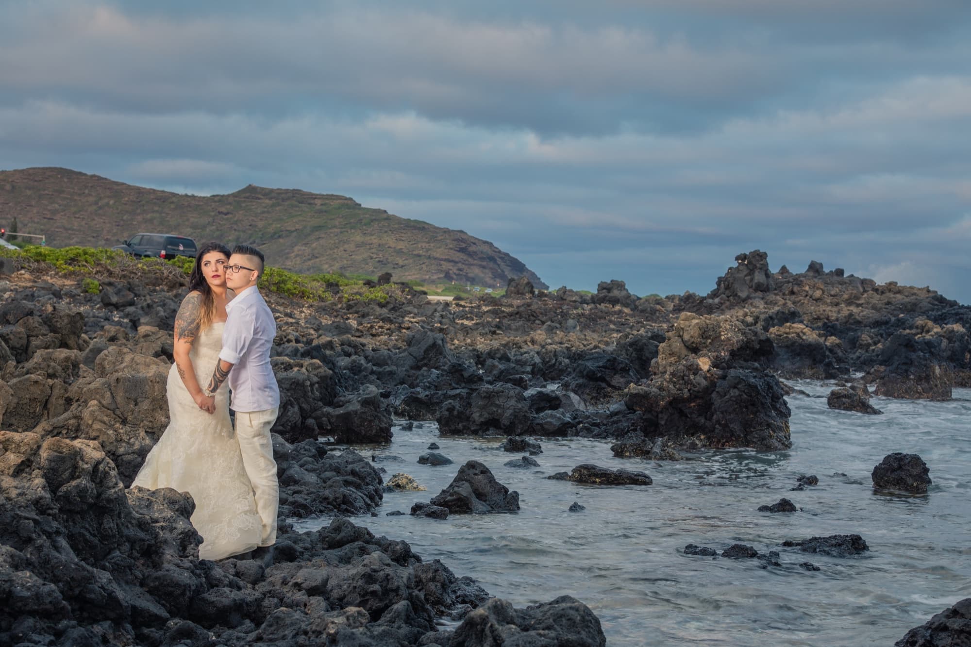 Amanda and Shauna embracing on the black lava rock coast of Oahu as the surf breaks behind them