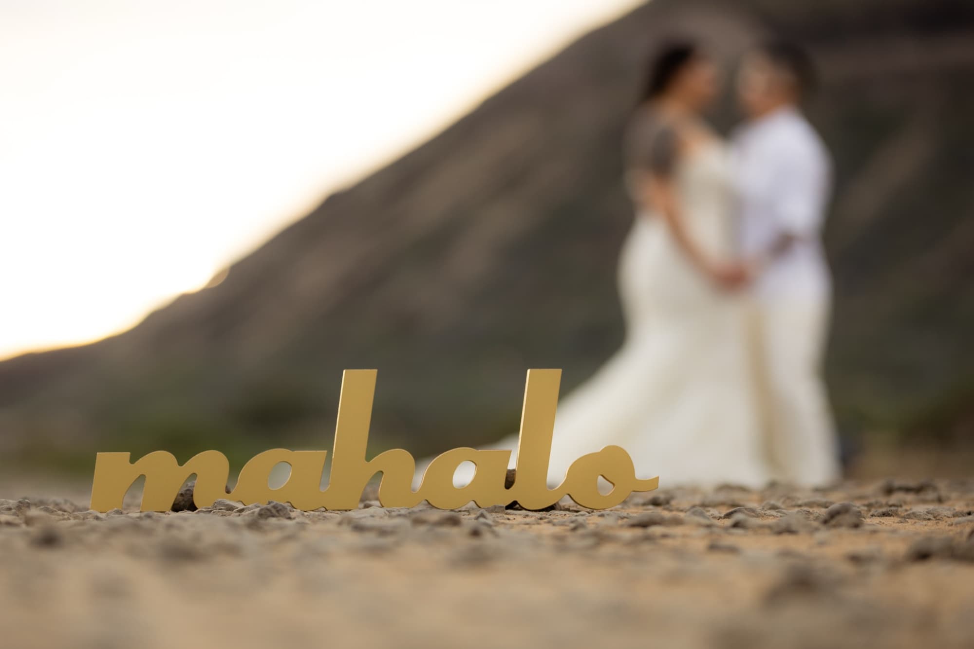 A gold mahalo sign in the sand with Amanda and Shauna embracing softly out of focus behind it at sunset