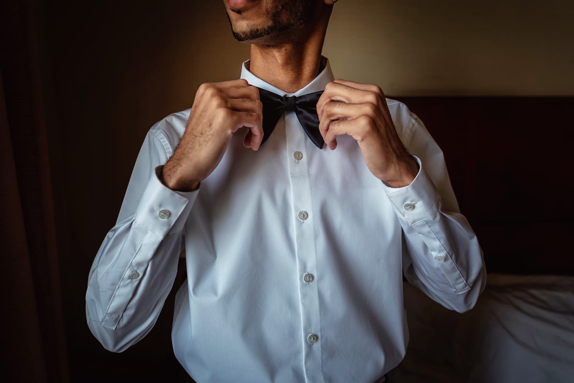 A groom in a crisp white shirt fastening his black bowtie during wedding prep at the Hilton Garden Inn Victorville