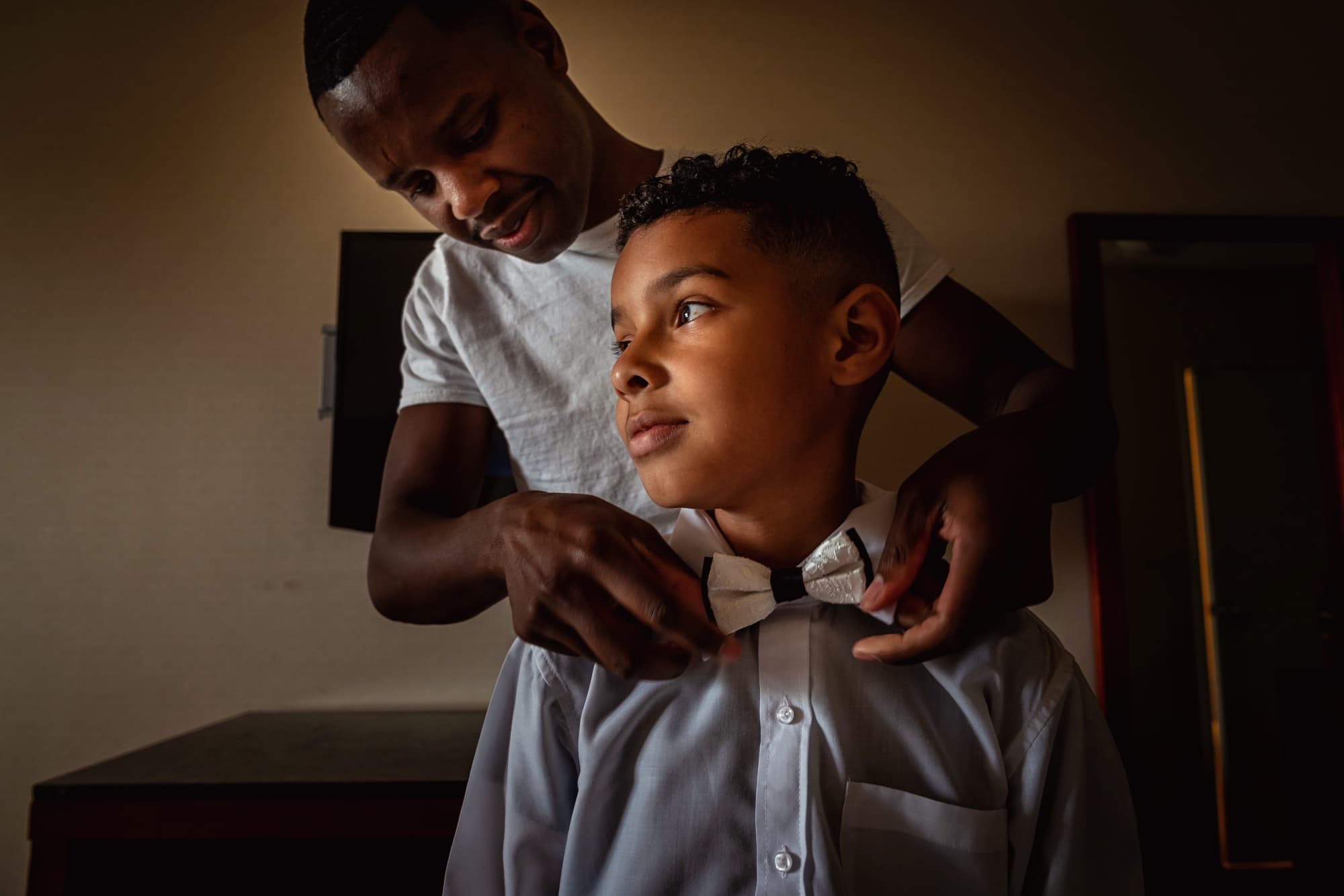 A young member of the wedding party getting his bowtie straightened over the shoulders in warm window light